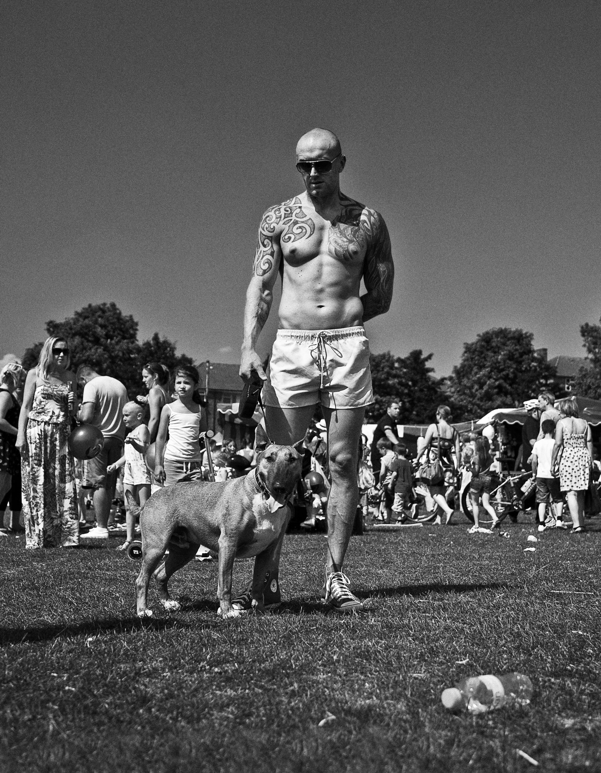 A shirtless man with tattoos, wearing sunglasses and shorts, walking a dog at an outdoor event with many people in the background.