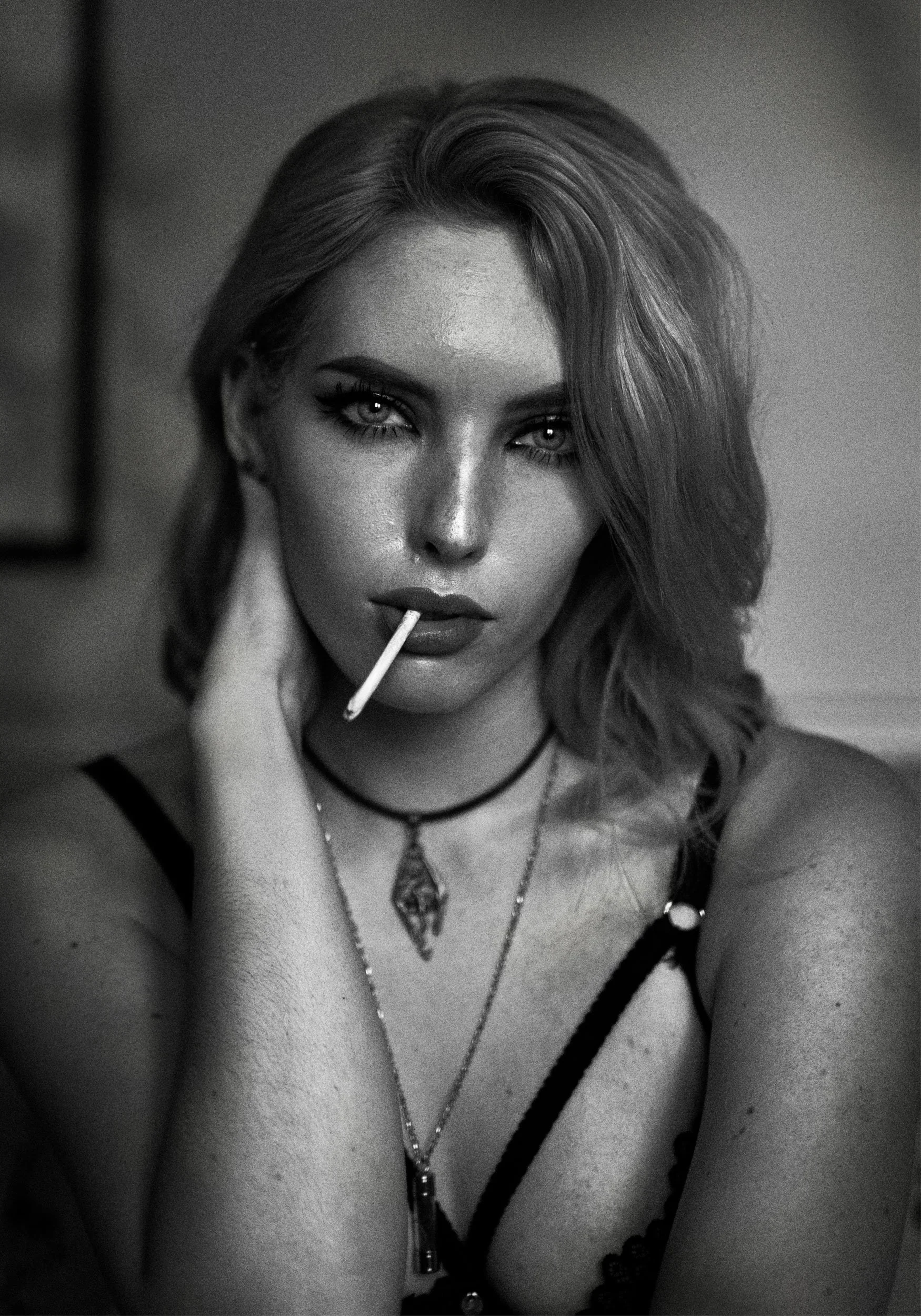 Black and white portrait of a woman with wavy hair, wearing a necklace, and holding a cigarette in her mouth.