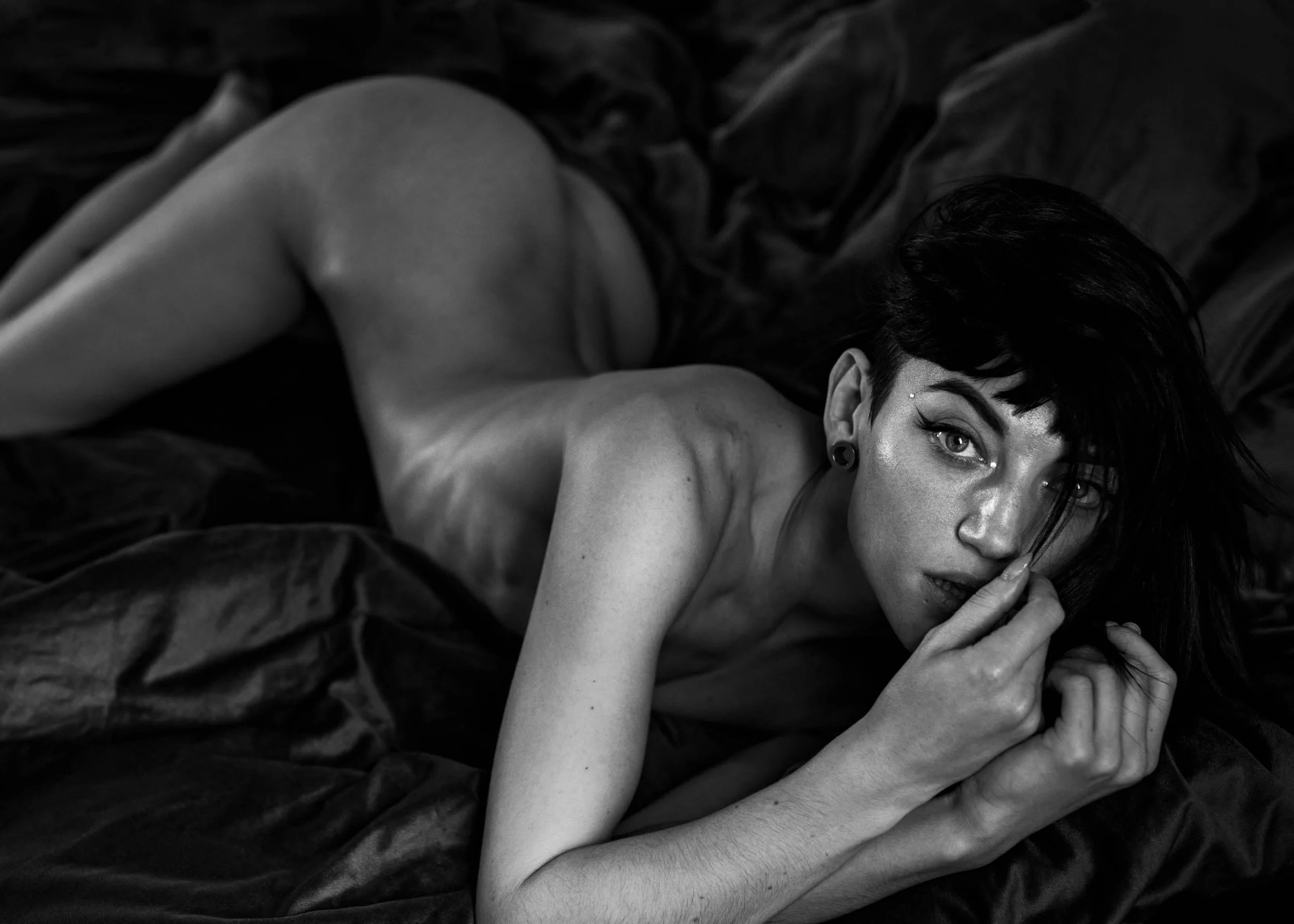 Black and white photo of a woman with short dark hair lying on a bed, looking at the camera, touching her face with her finger.