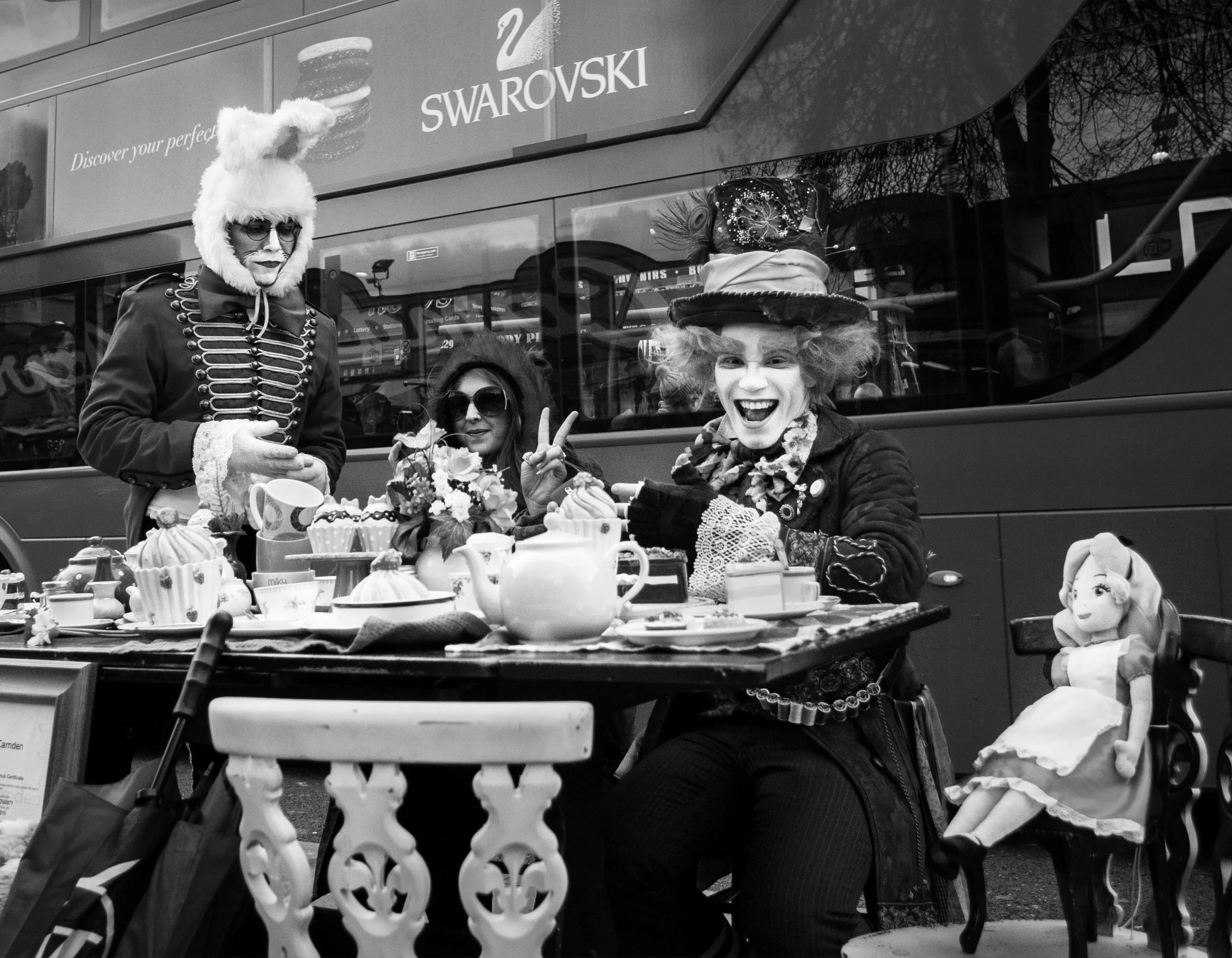 People dressed as characters at a table with tea and cakes in front of a bus with Swarovski advertisement in the background.