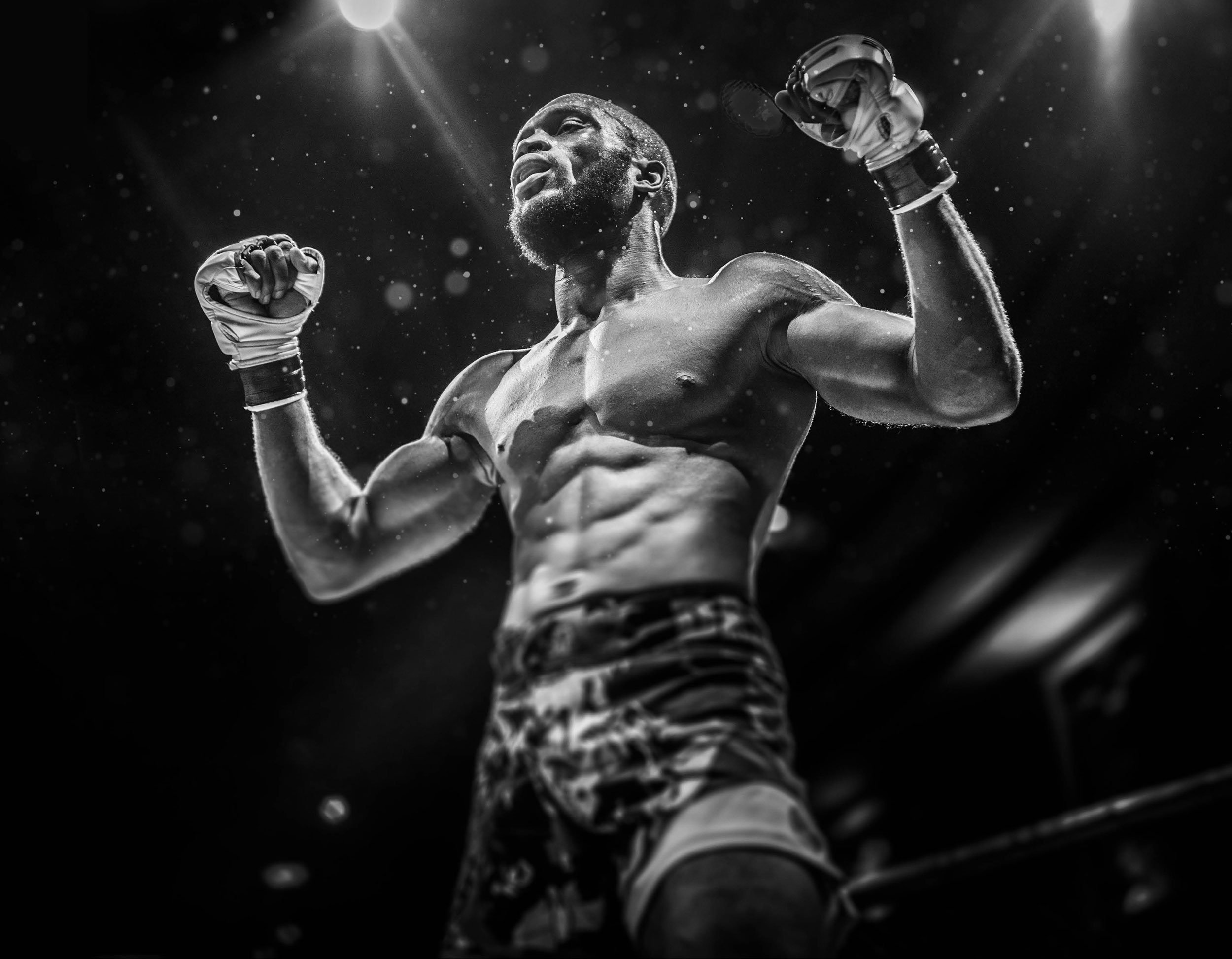 Black and white photo of a male boxer flexing muscles in a fighting stance, wearing boxing gloves and shorts, with a spotlight shining from above.