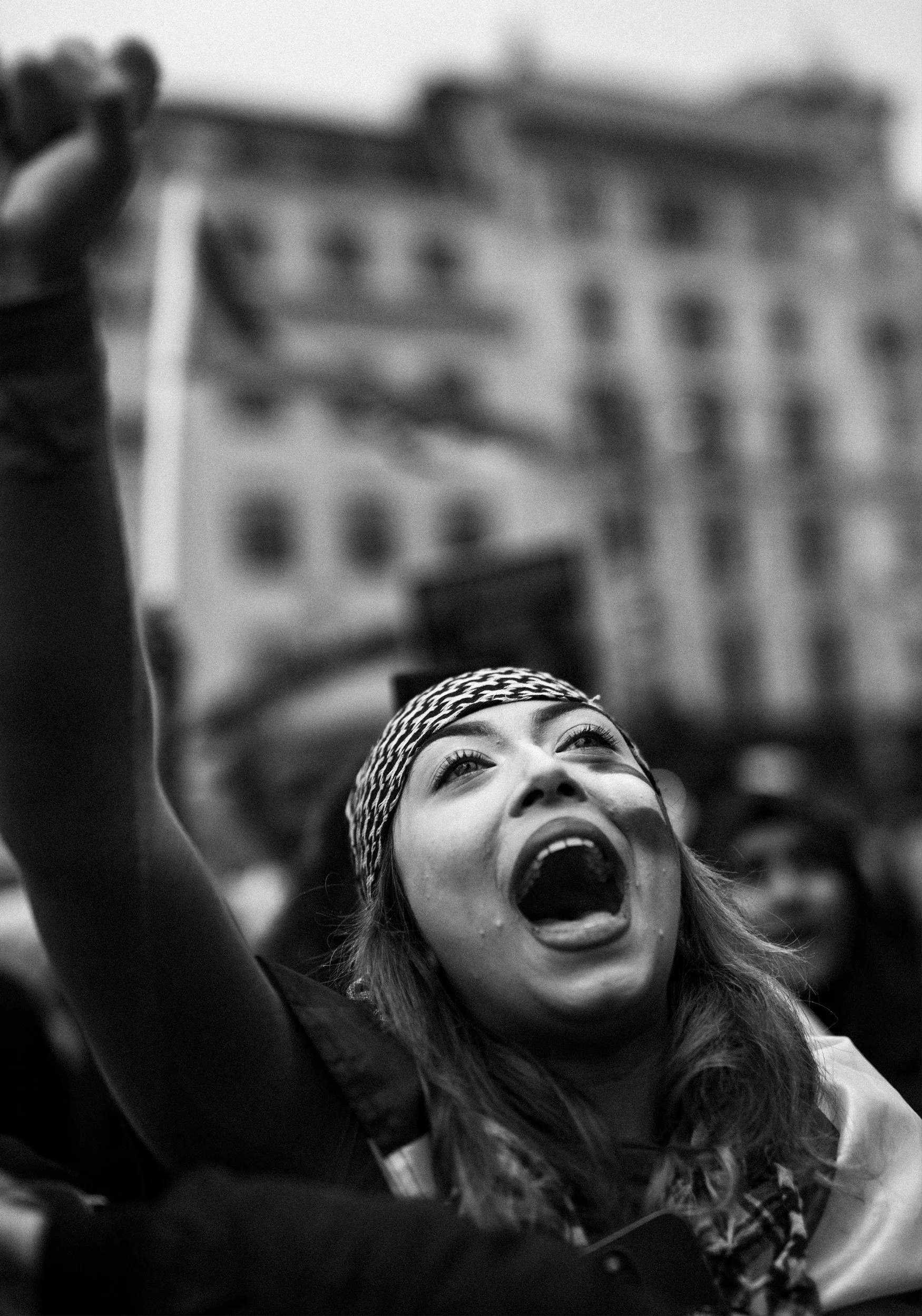A woman with long hair and a headband is shouting with her mouth open and her eyes wide, raising her arm in the air. She appears to be participating in a protest or rally, with a blurred building in the background.