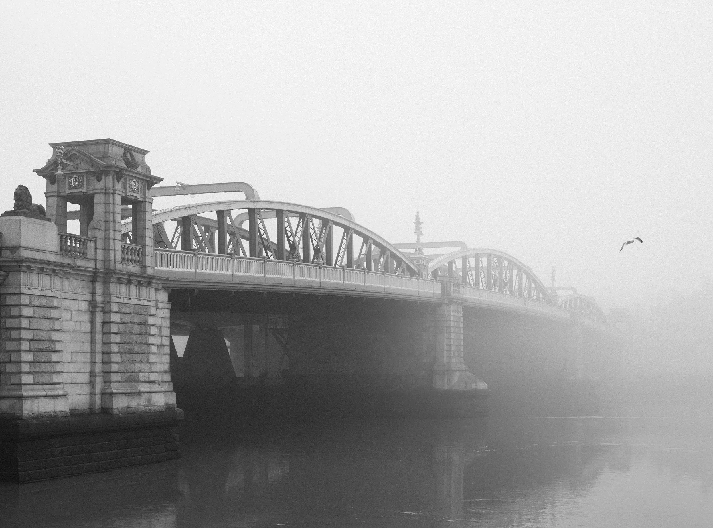 Black and white photo of a foggy river with a historic stone bridge featuring arches and decorative elements, and a lone bird flying nearby. Rochester bridge 