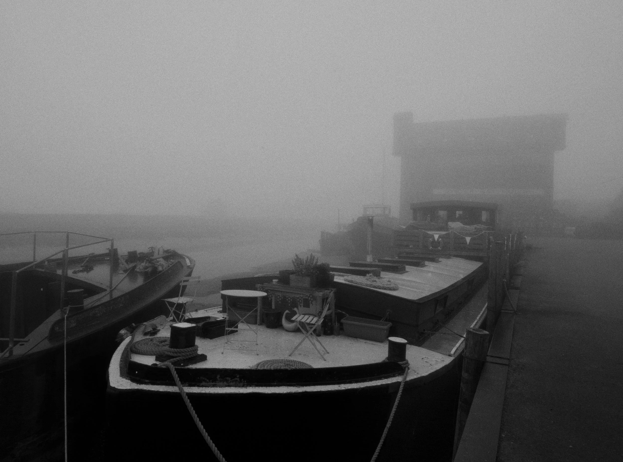 Black and white photo of boats docked at a foggy waterfront, with a wooden pier and a building in the background.