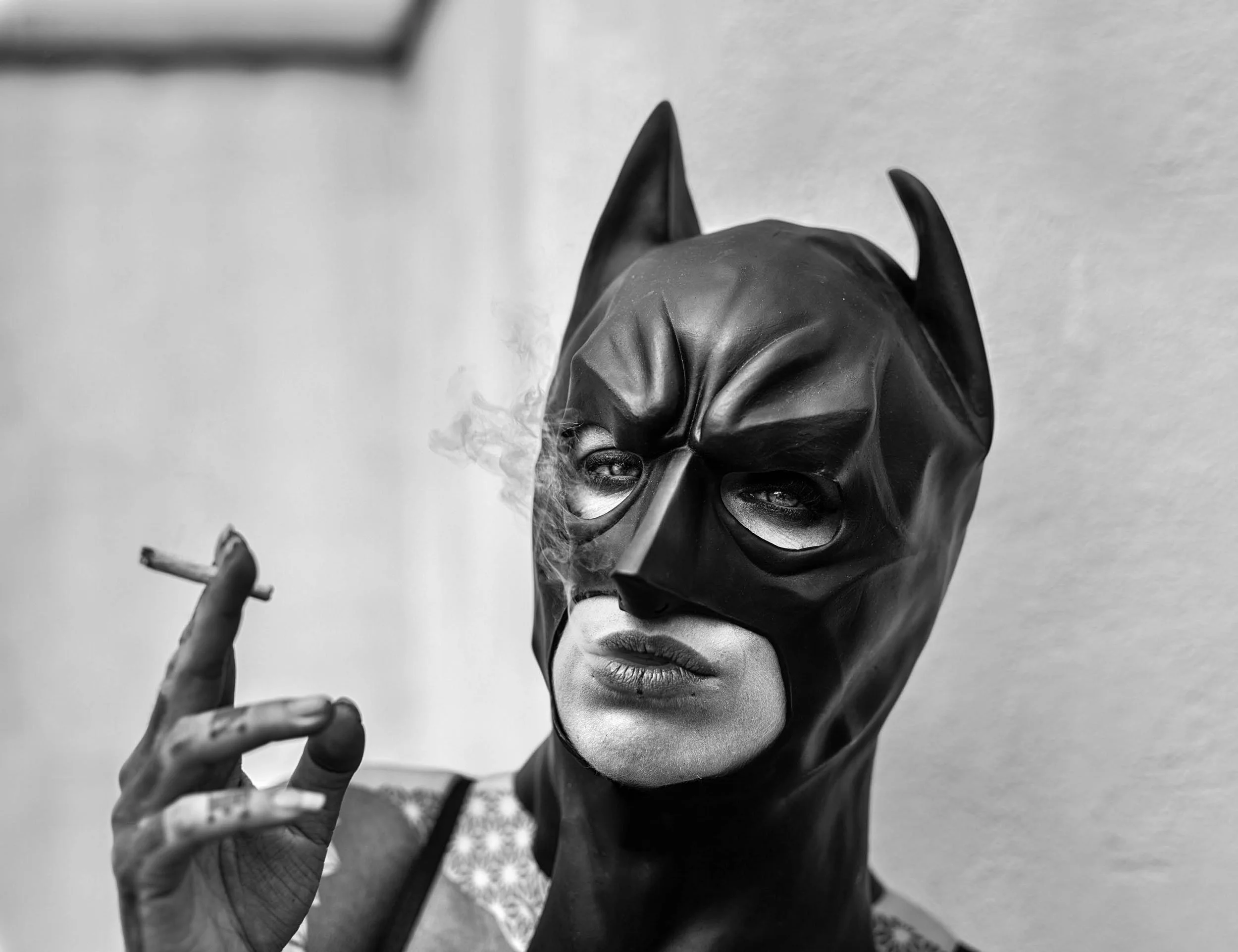 Black and white photograph of a woman wearing a leather mask with cat ears and reading glasses, making a peace sign with her hand.