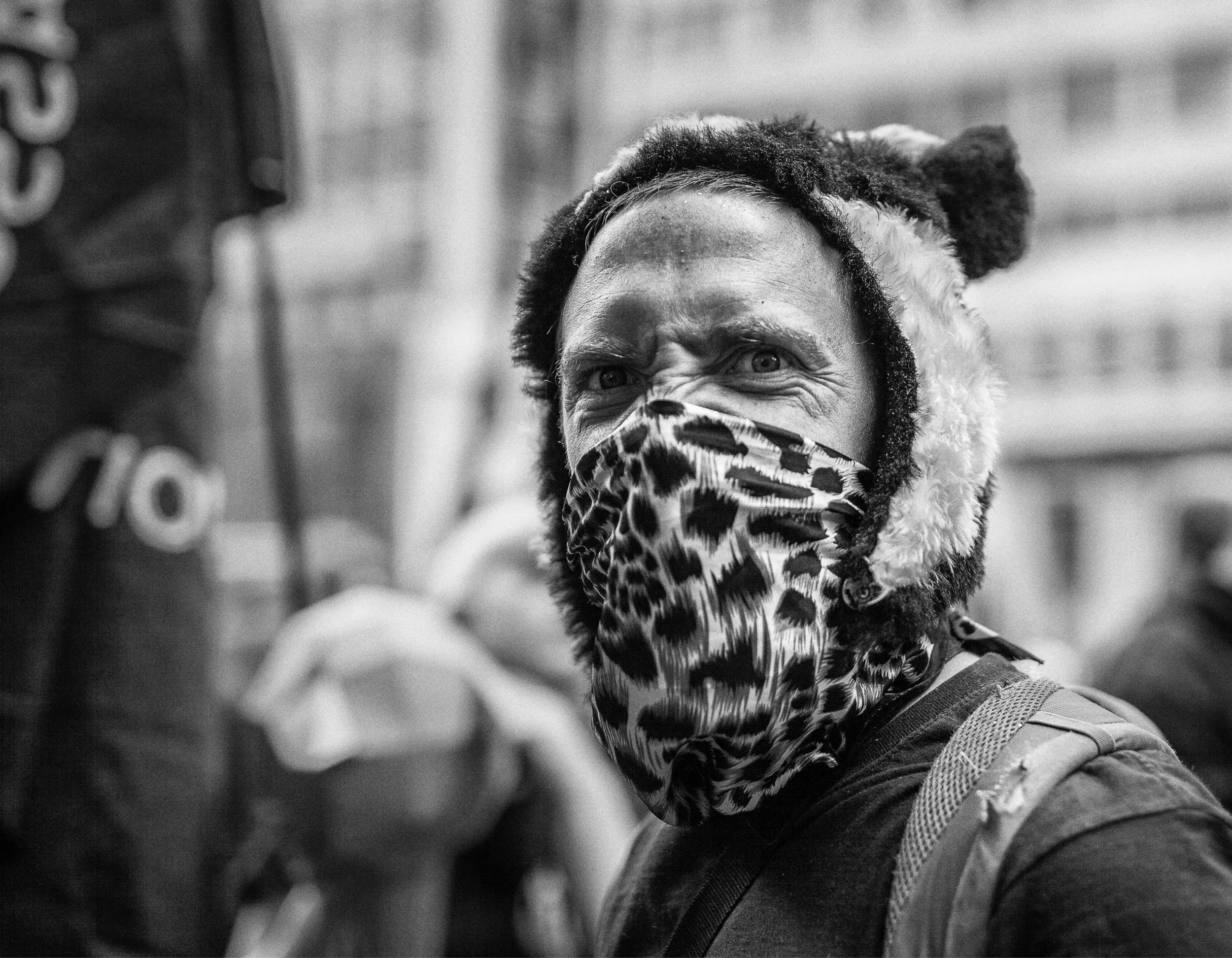 A person with wrapped dreadlocks wearing a leopard print face mask and hoodie, looking intensely at the camera on a city street.