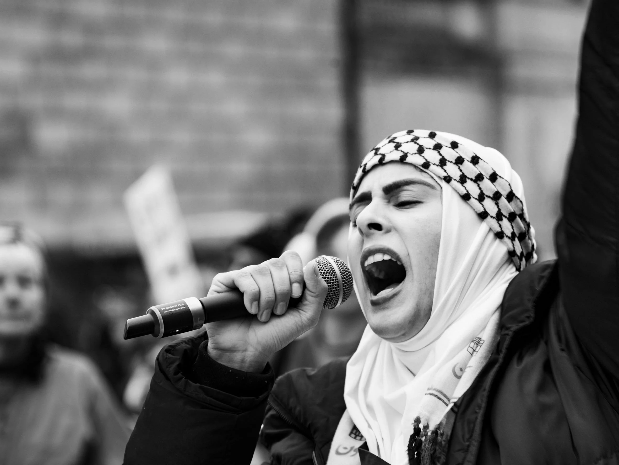 A person wearing a keffiyeh and jacket passionately speaking or shouting into a microphone during a protest or rally, with a blurred crowd in the background.