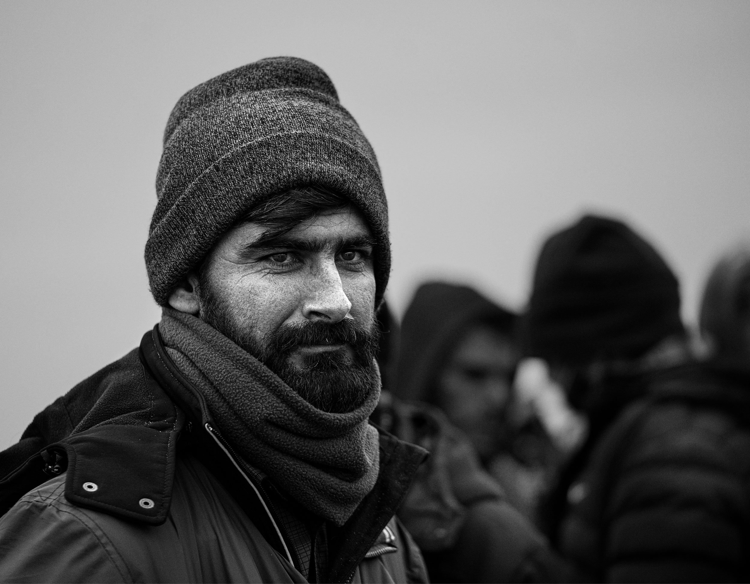 A black and white photo of a man with a beard and mustache wearing a knit cap and a scarf, while other people are blurred in the background.