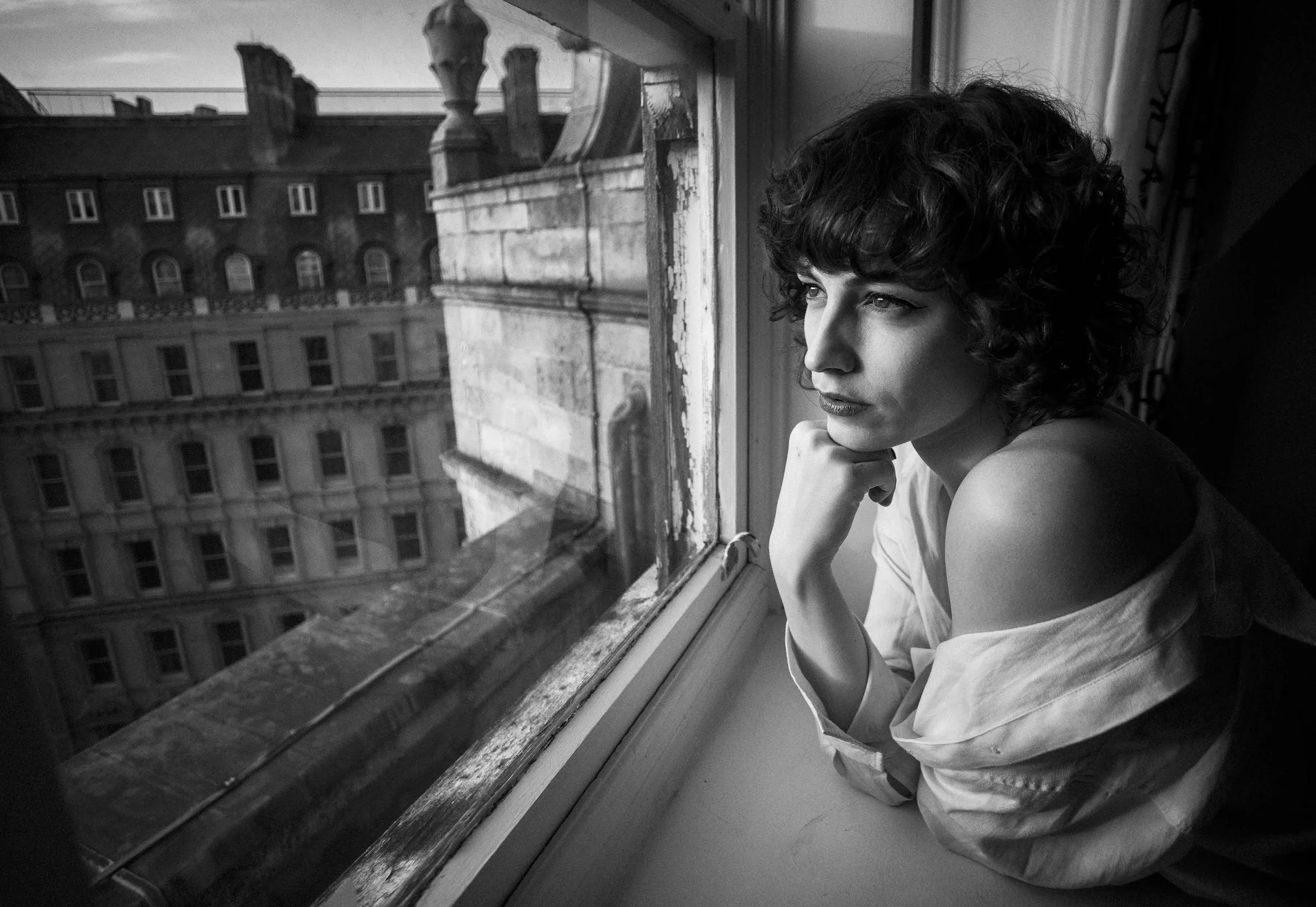A woman with curly hair looking out of a window on a building's rooftop, with her chin resting on her hand, in black and white.