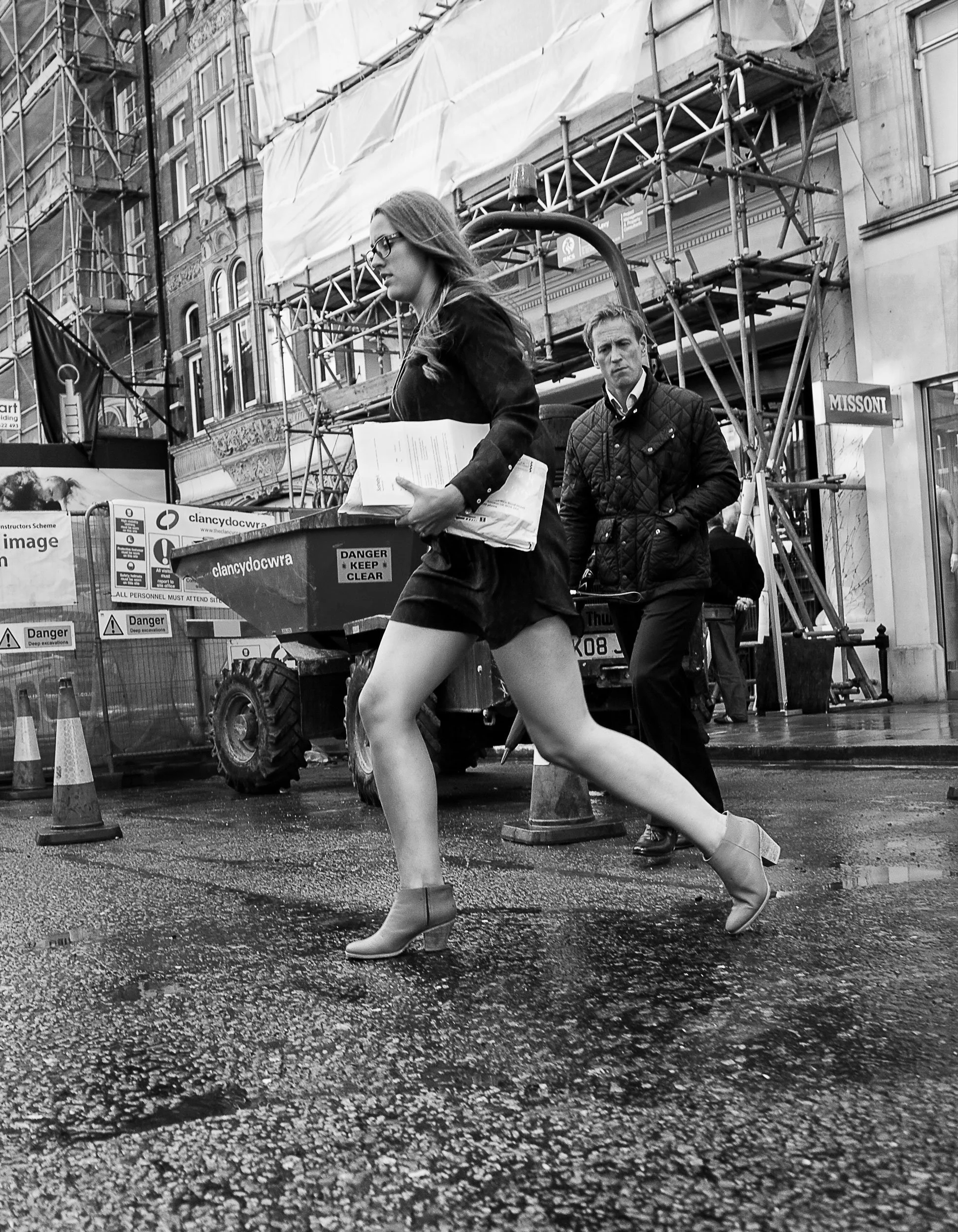 A woman wearing glasses, a dark jacket, and ankle boots walking on a wet city street while holding papers and a box, with a man in a dark quilted jacket behind her. Construction scaffolding and barriers are visible in the background.