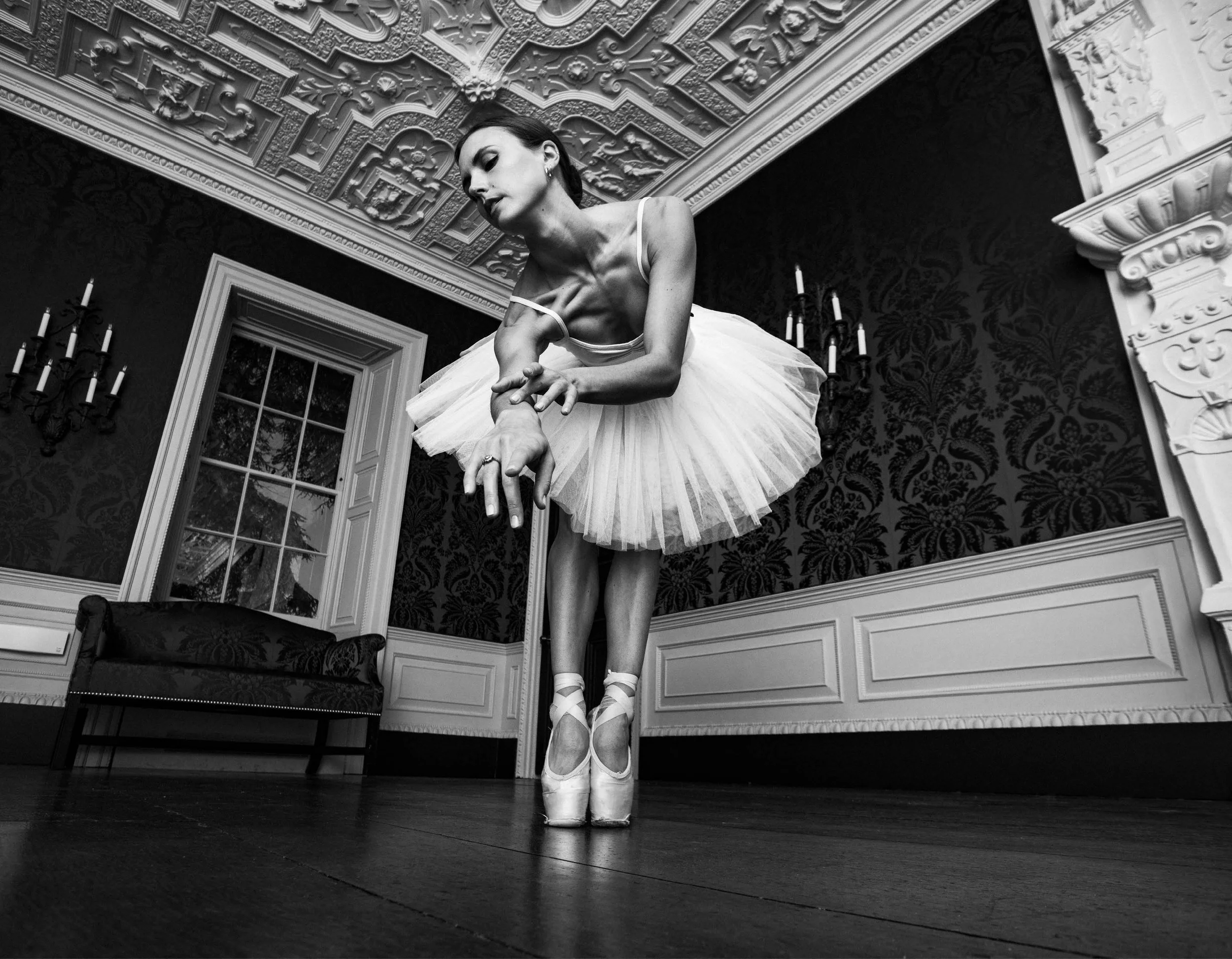 Black and white photo of a ballerina on pointe, wearing a tutu, dancing in an ornate room with decorative ceiling, chandelier, and wainscoting.