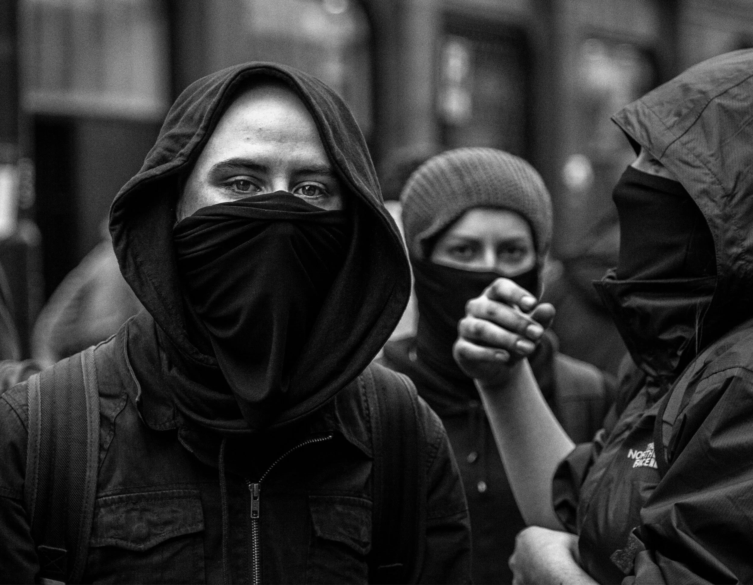Three people, two men and one woman, wearing black masks covering their noses and mouths, dressed warmly with hooded jackets, gathered in an urban setting.