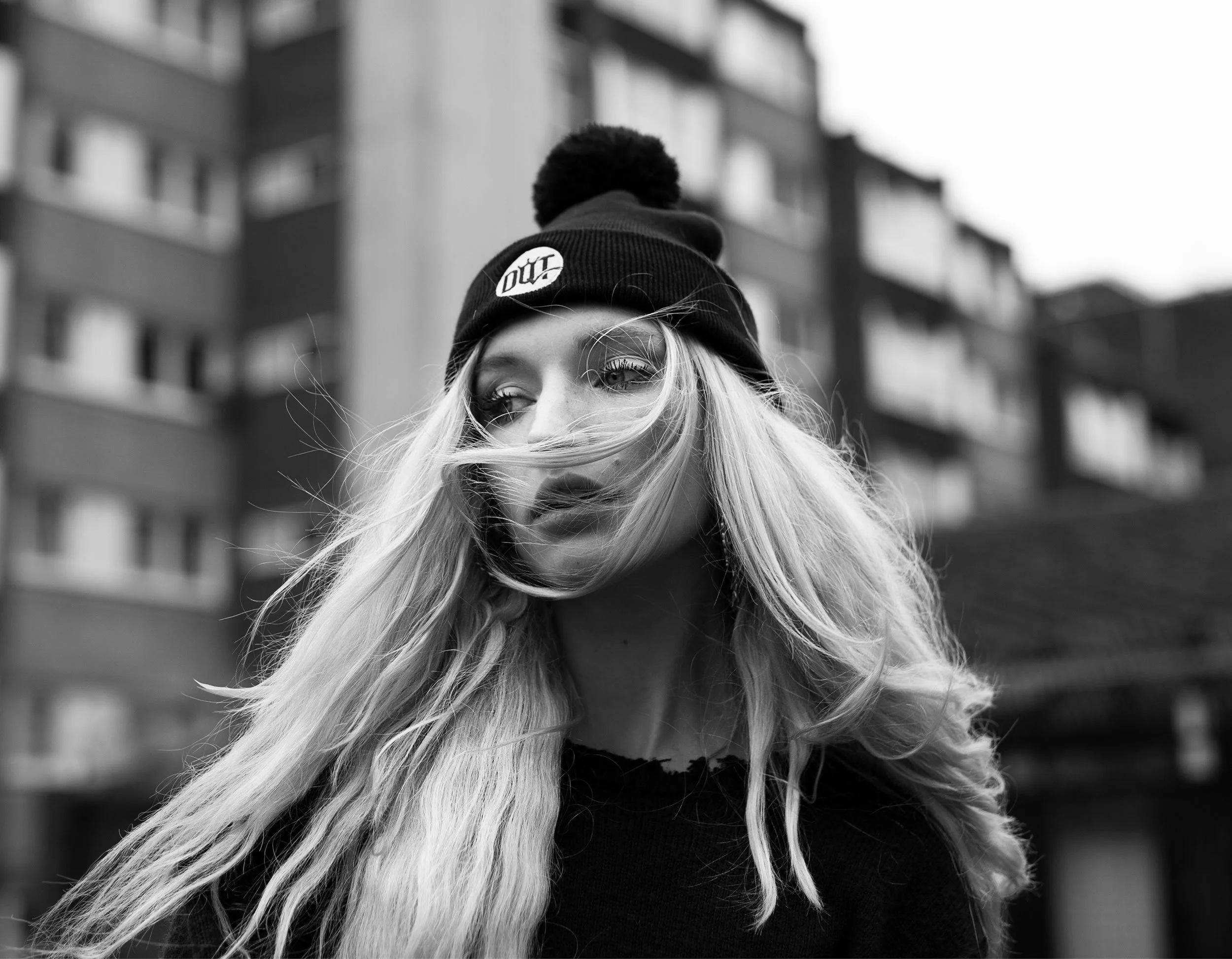 Black-and-white photo of a young woman with long wavy hair, wearing a beanie hat, standing outdoors in front of a multi-story building, with wind blowing her hair across her face.