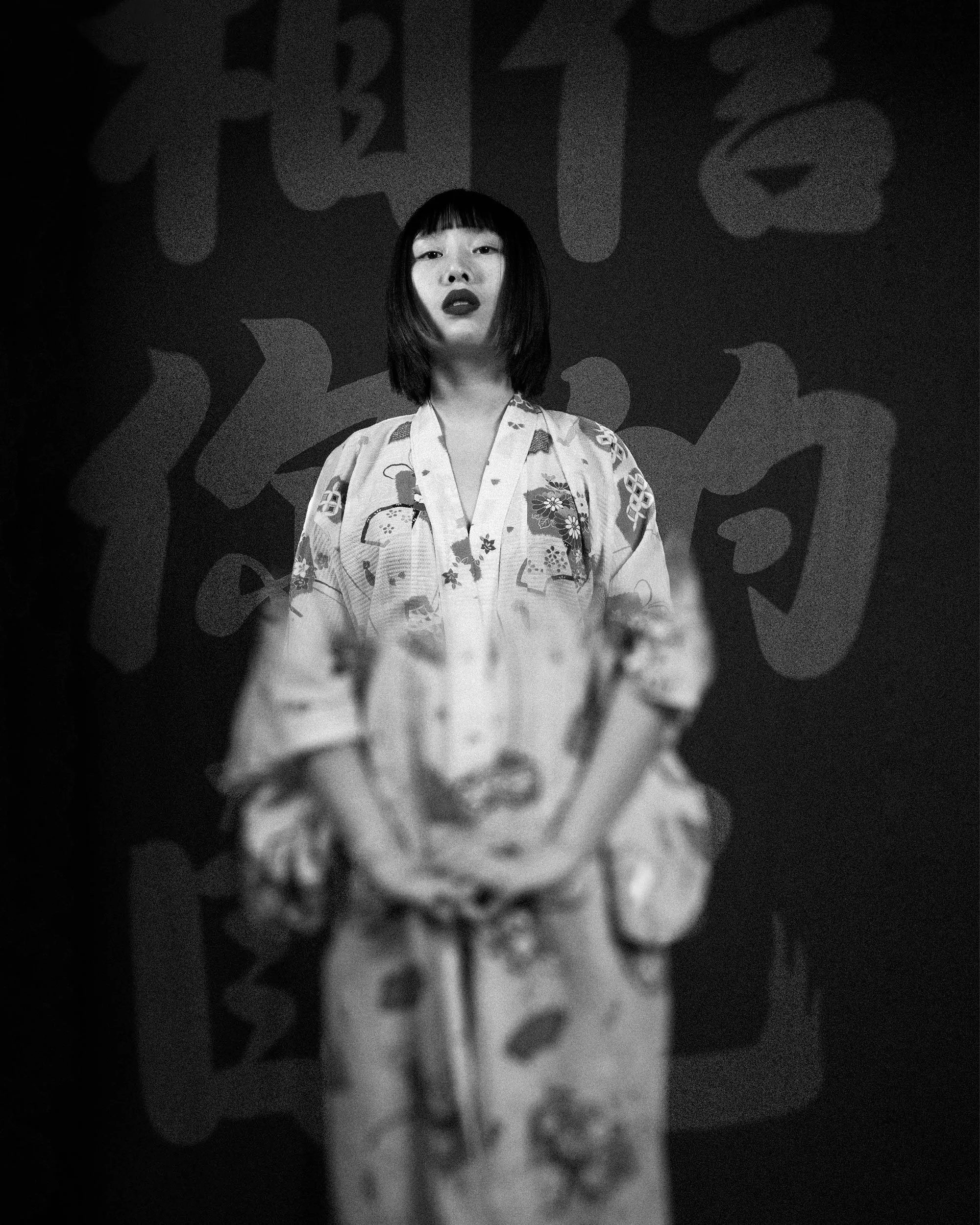 A woman with shoulder-length dark hair in a kimono standing against a backdrop with large Japanese characters.