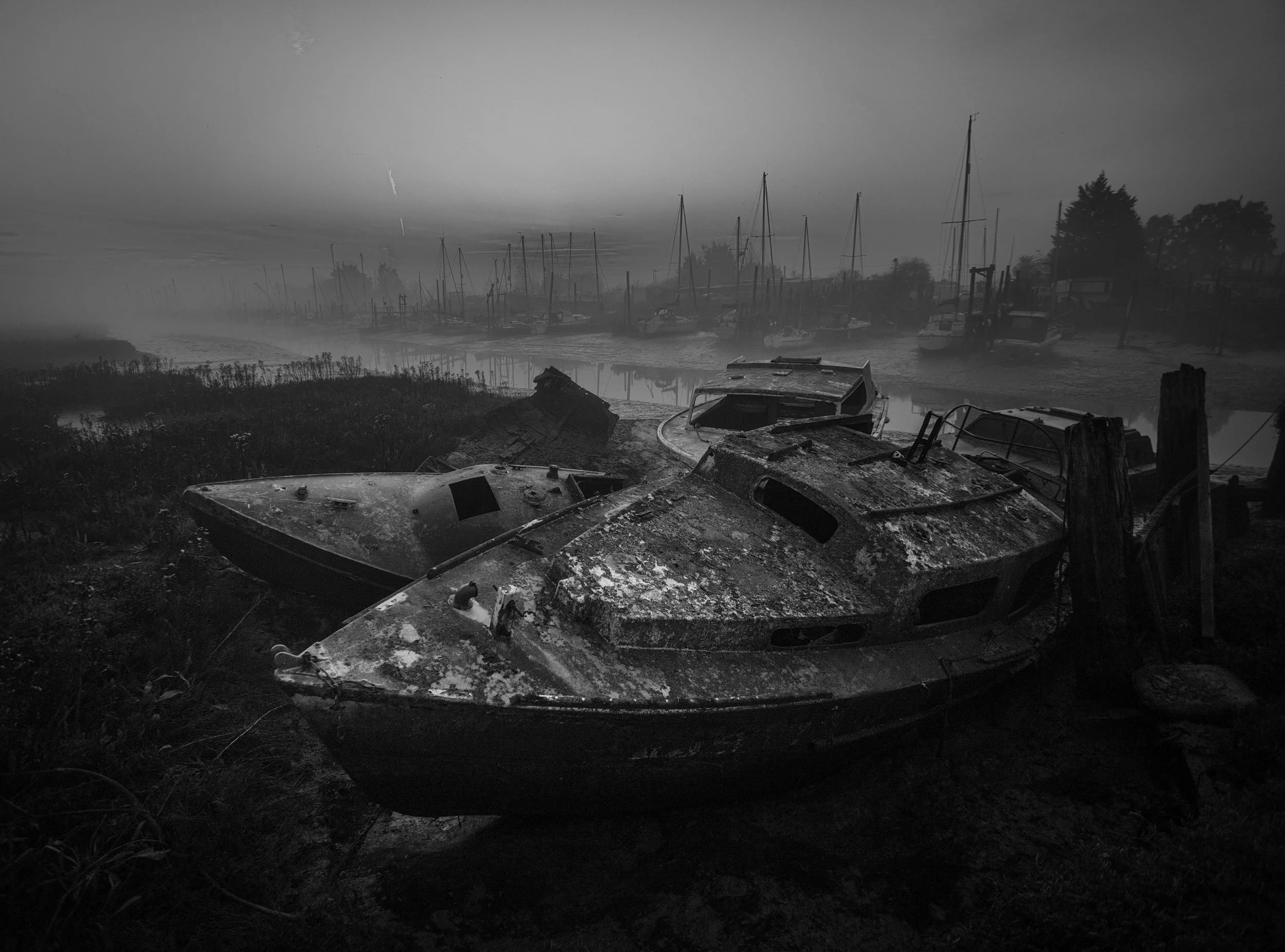 Black and white photograph of abandoned, rusted boats on a foggy shoreline with a marina and sailboats in the background.