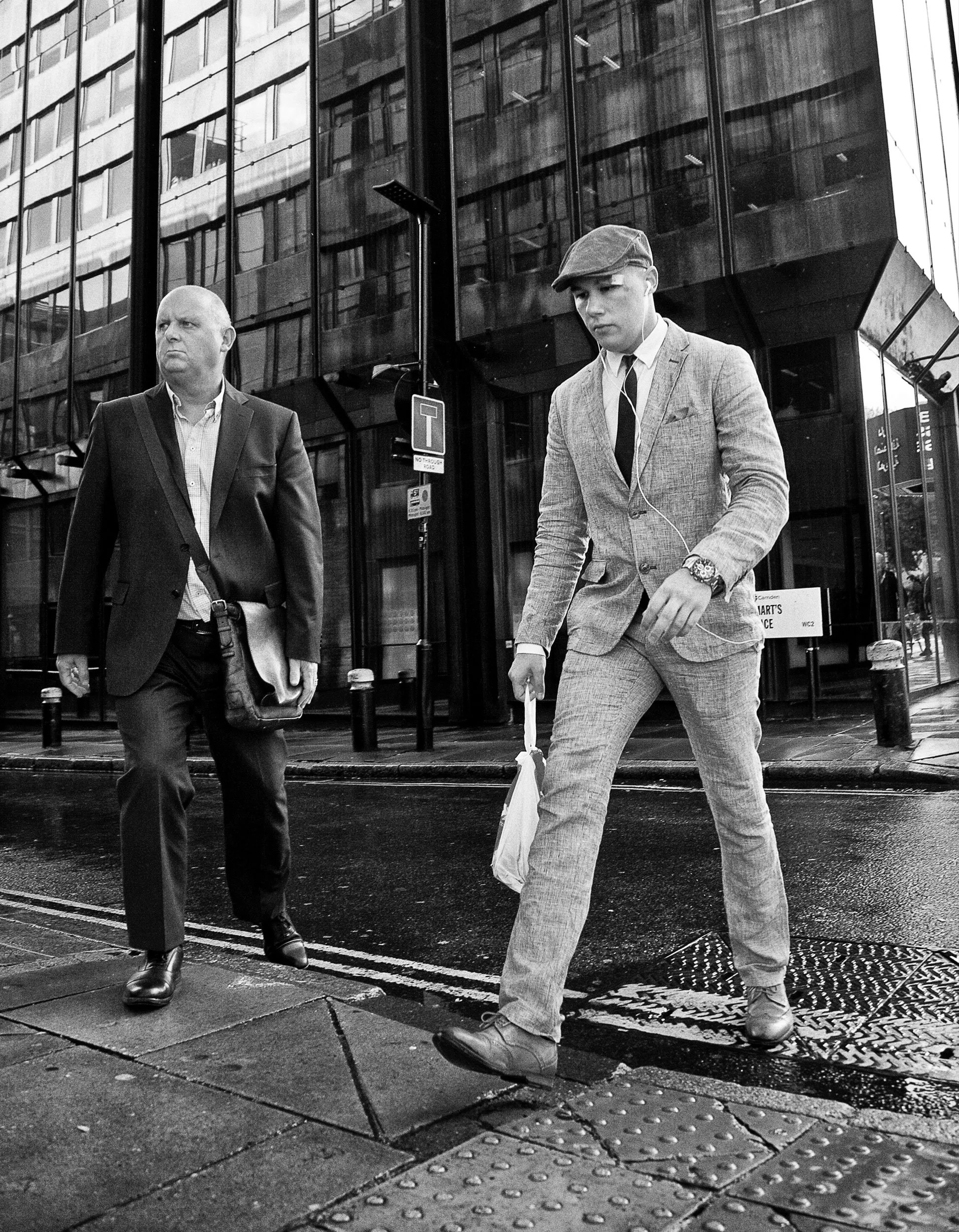 Black and white photo of two men walking on a city sidewalk. One man is bald, wearing a suit with a bag, and looking serious. The other man is younger, wearing a light-colored suit and flat cap, carrying a shopping bag, with earphones.