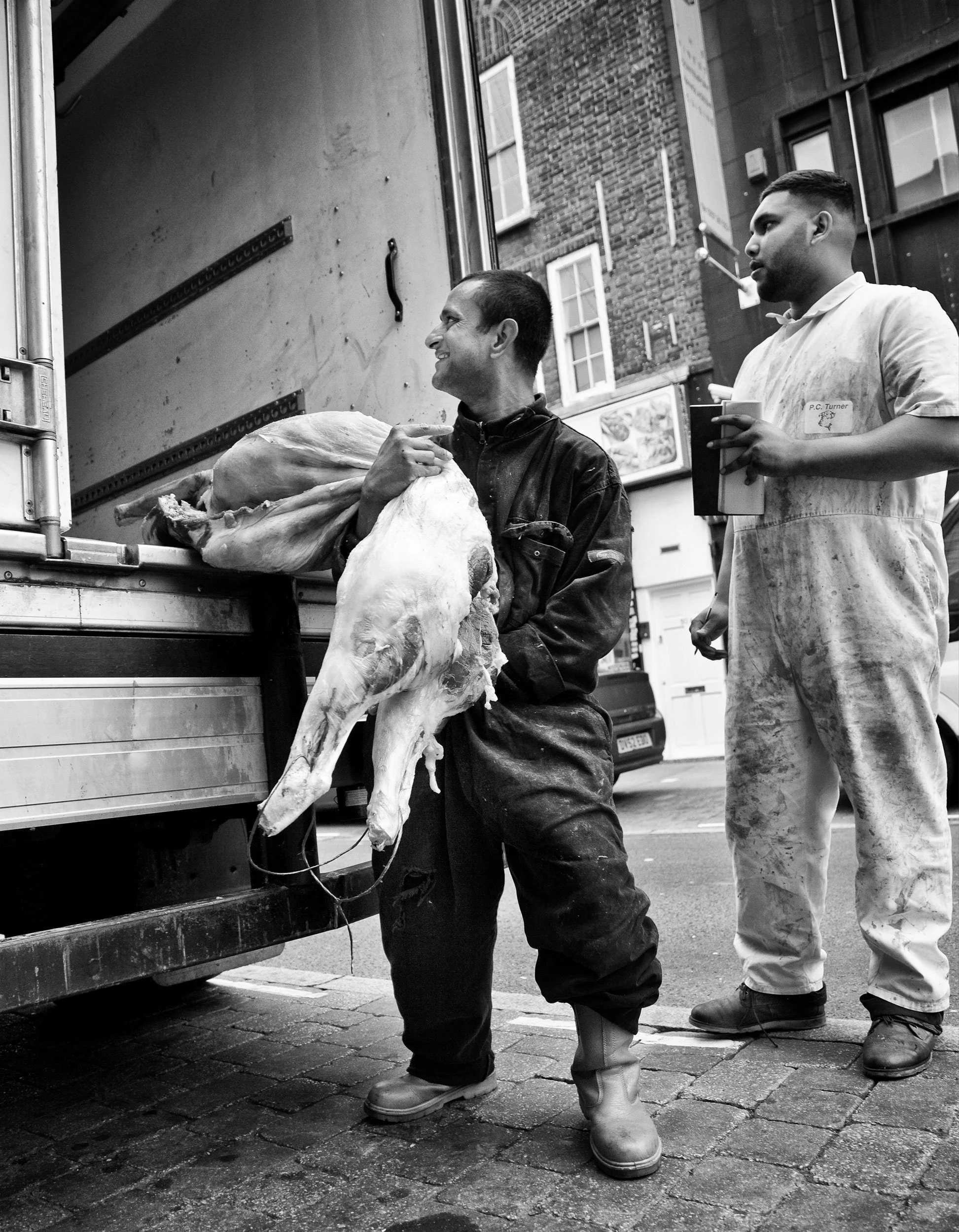Two men, one in a butcher's apron, standing next to a food truck with a carcass of a sheep or goat, on a city street.