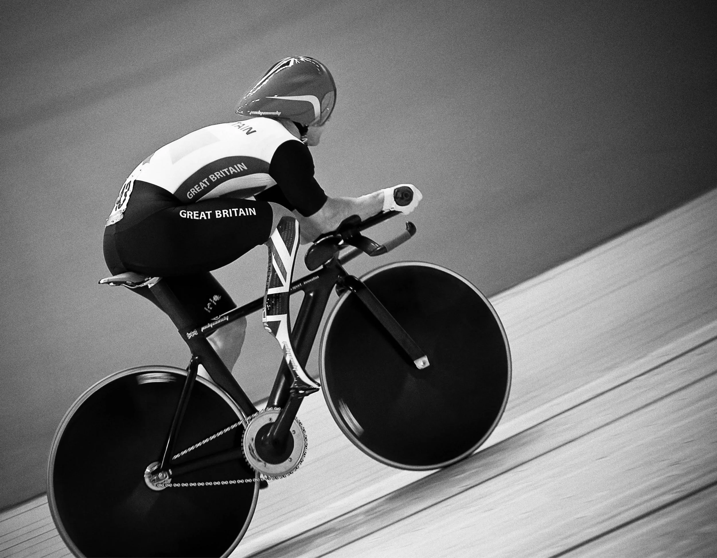 A cyclist in a Great Britain uniform and helmet riding a track bike on an indoor velodrome with a wooden track, in black and white.