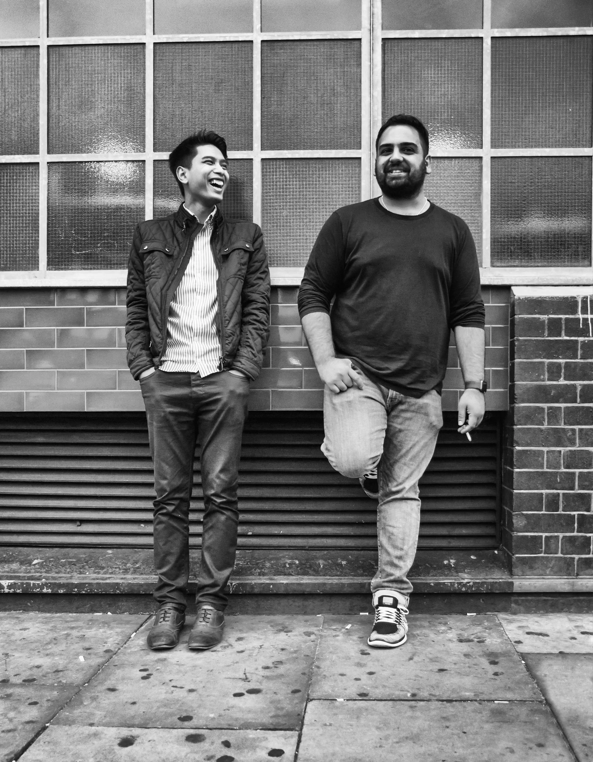 Two men standing outside in front of a brick wall and large windows, one laughing and the other smiling, dressed casually.