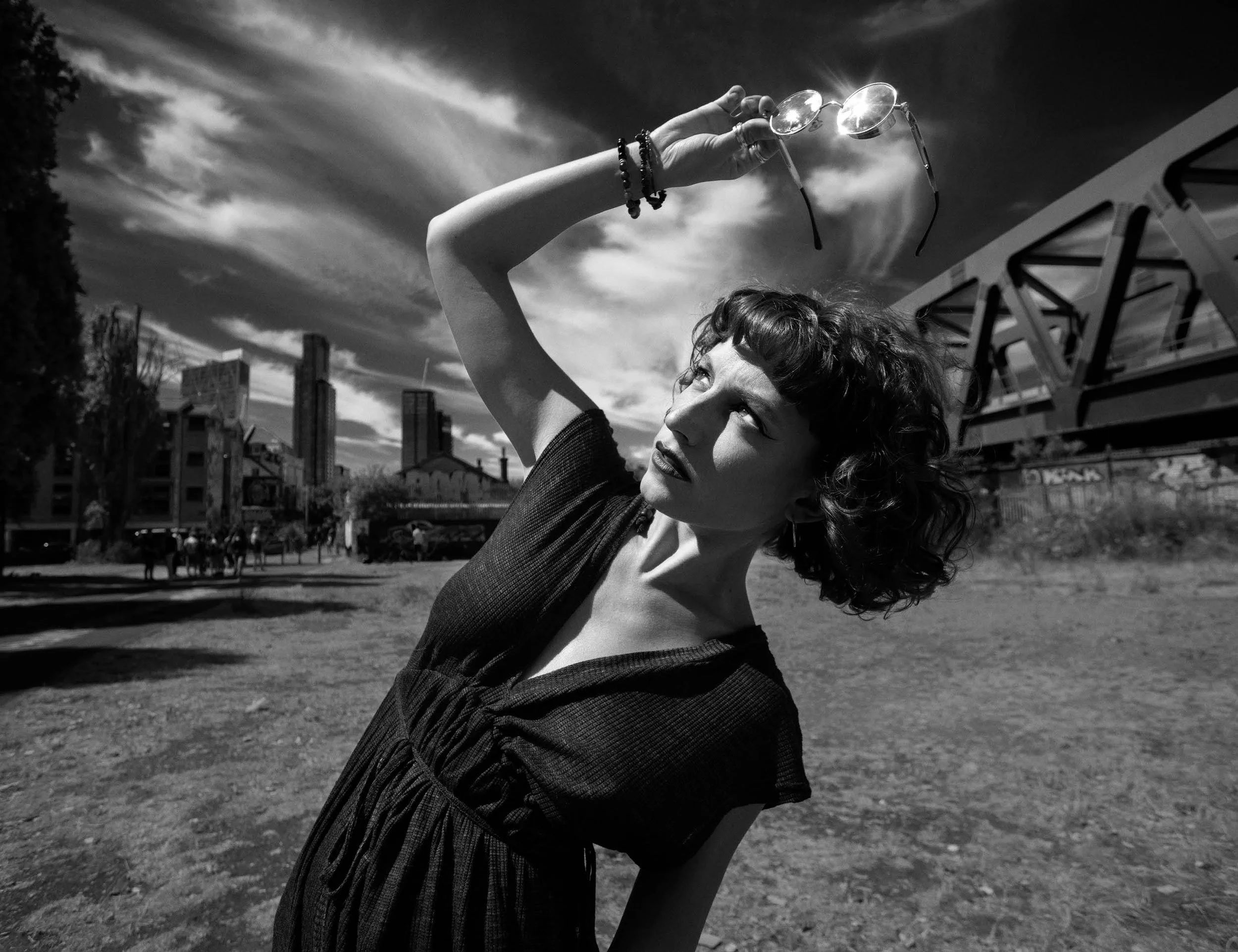 A woman with curly hair holding sunglasses high above her head outdoors in an urban park, with city buildings and a partly cloudy sky in the background.