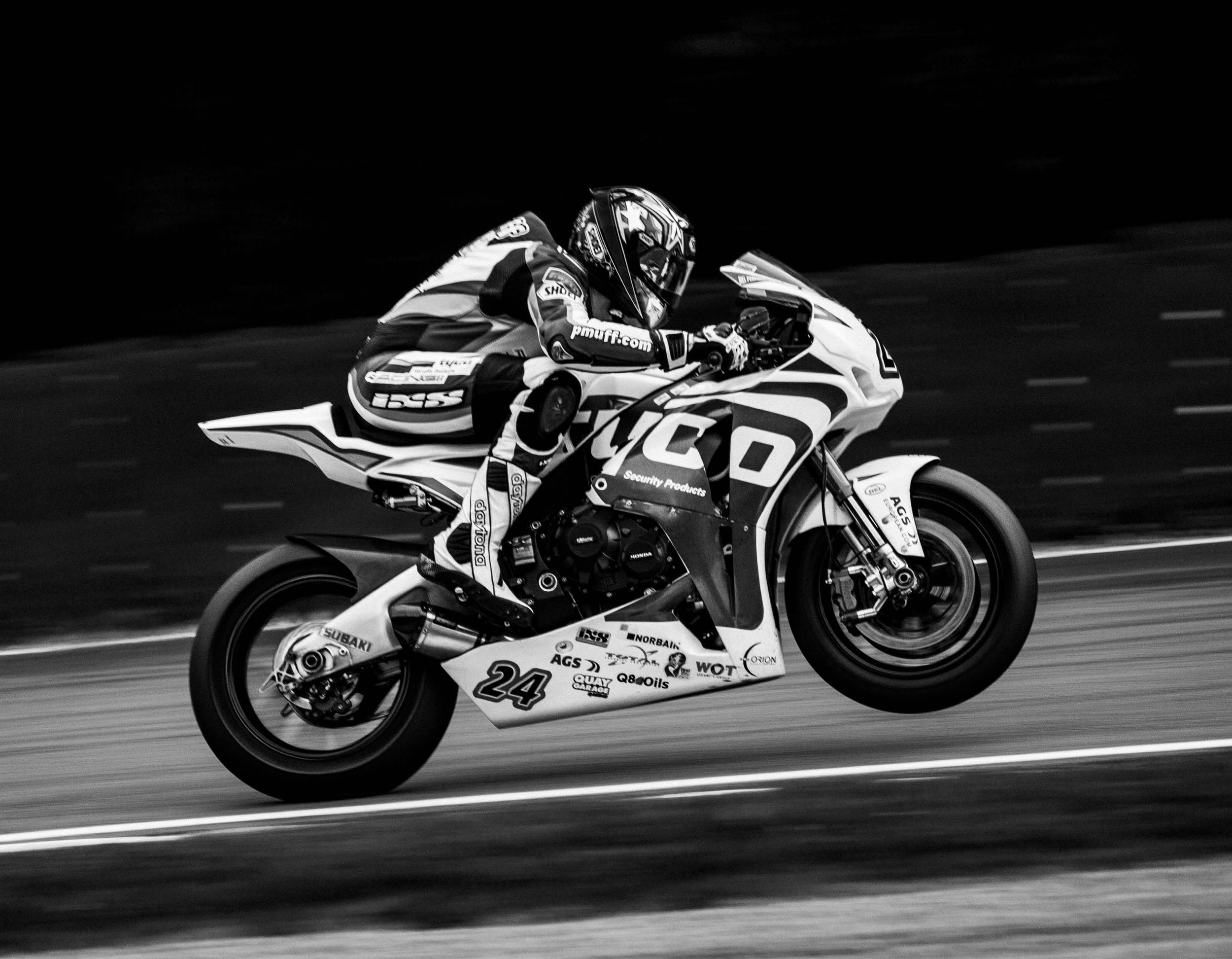 A motorcyclist wearing a helmet and racing gear riding a high-performance race bike on a track, black and white photo with motion blur.