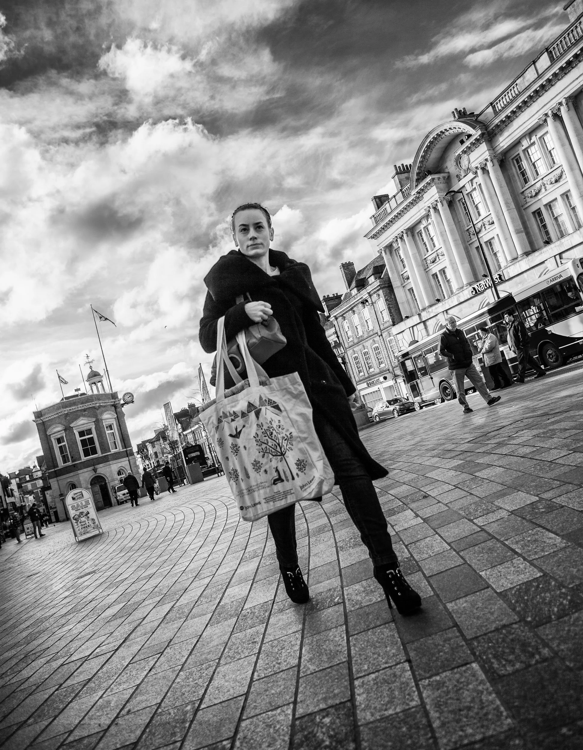 A woman in a black coat and high-heeled boots walking on a cobblestone street, carrying a reusable shopping bag and a purse, with historic buildings and a bus in the background under a cloudy sky.