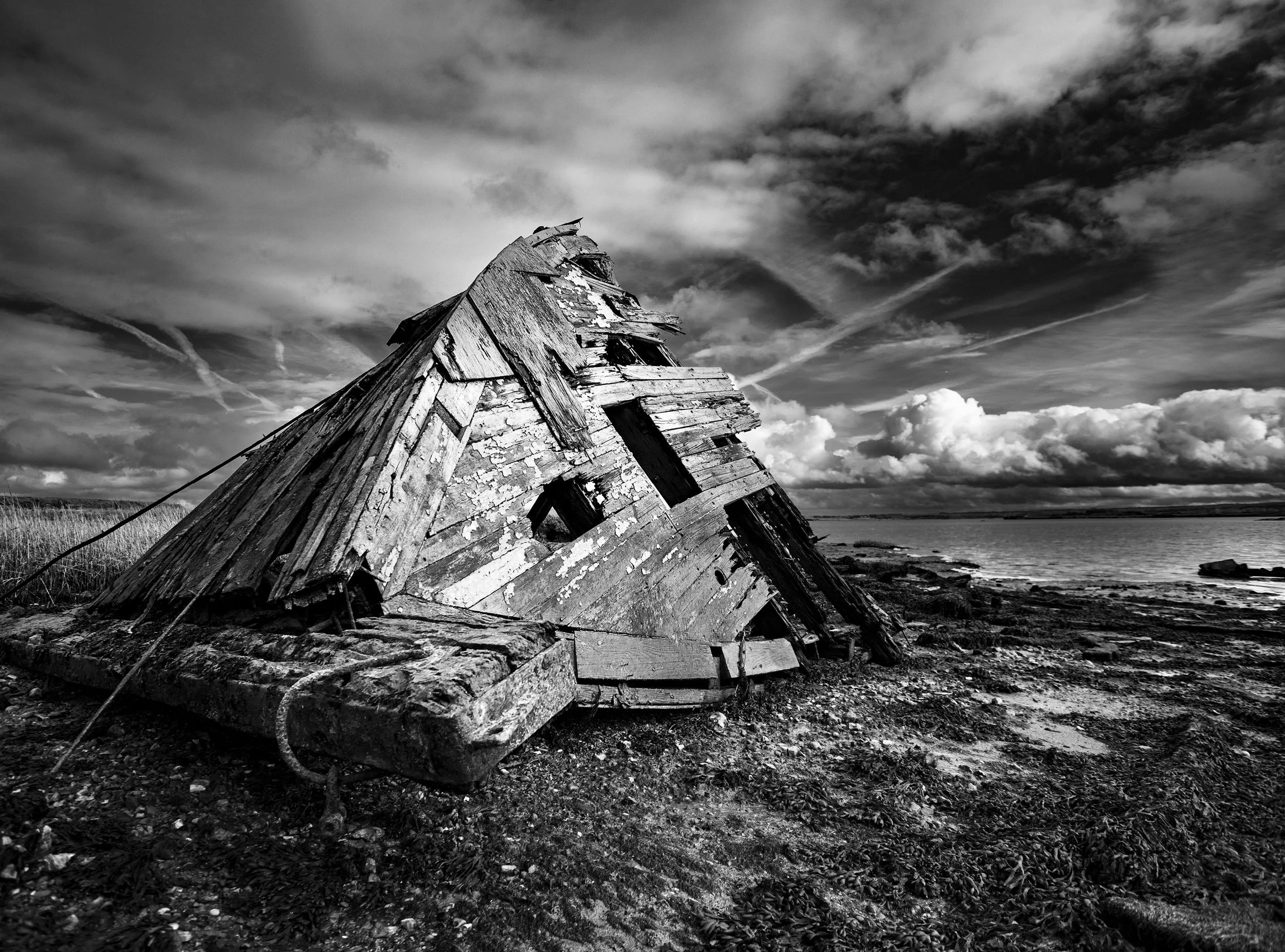 Black and white photo of a dilapidated, leaning wooden boat on a rocky shoreline, with cloudy sky and water in the background. Derelict boat in Hoo, Rochester, UK