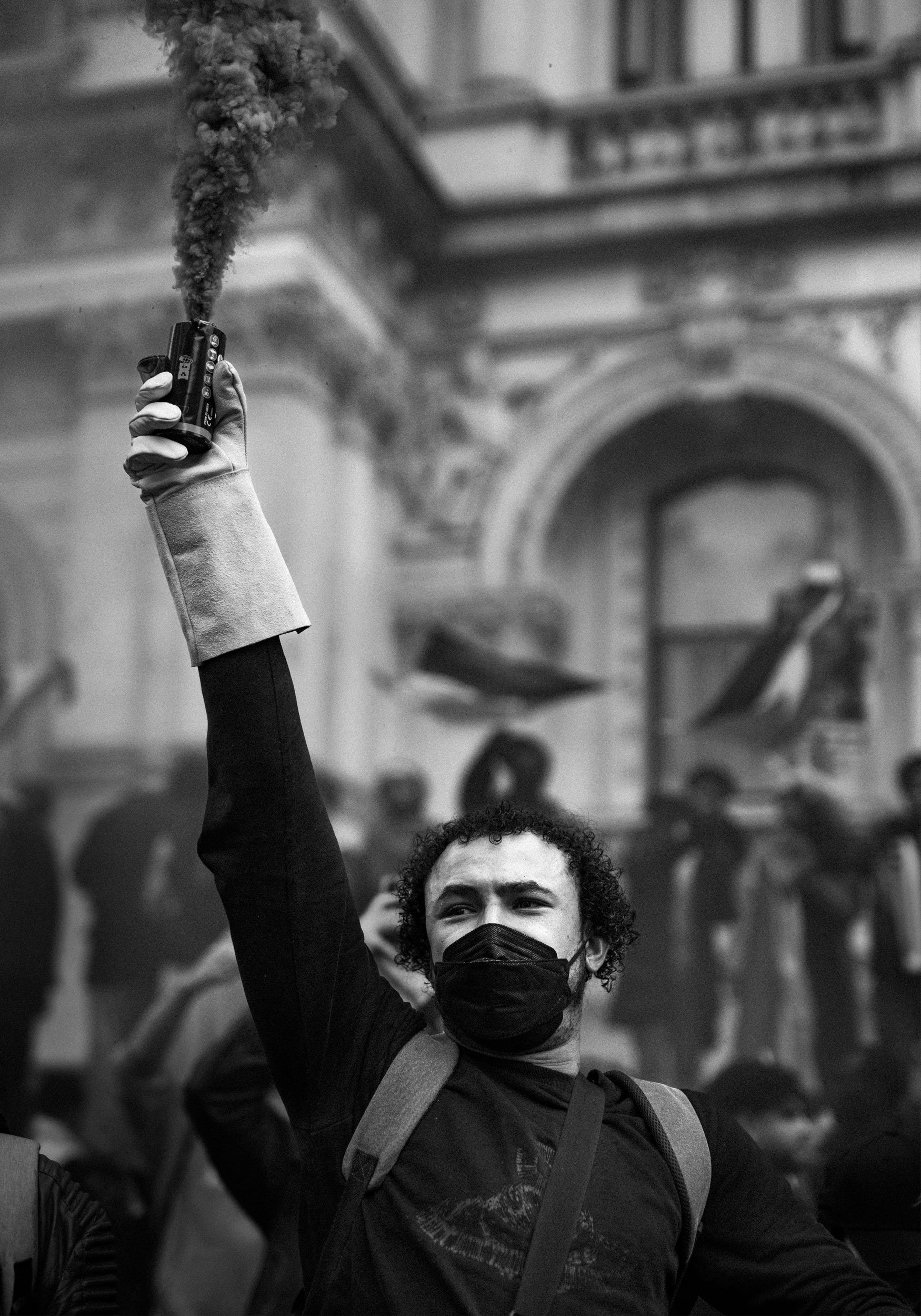 A young man with curly hair and a black face mask holding a smoke bomb or flare in a protest, with a crowd and classical building in background.