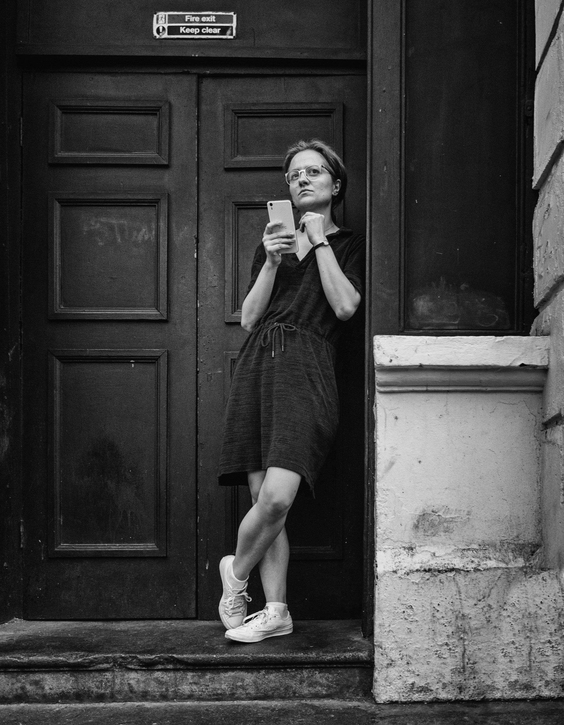 A woman standing against a wooden door, holding a smartphone and looking to the side.