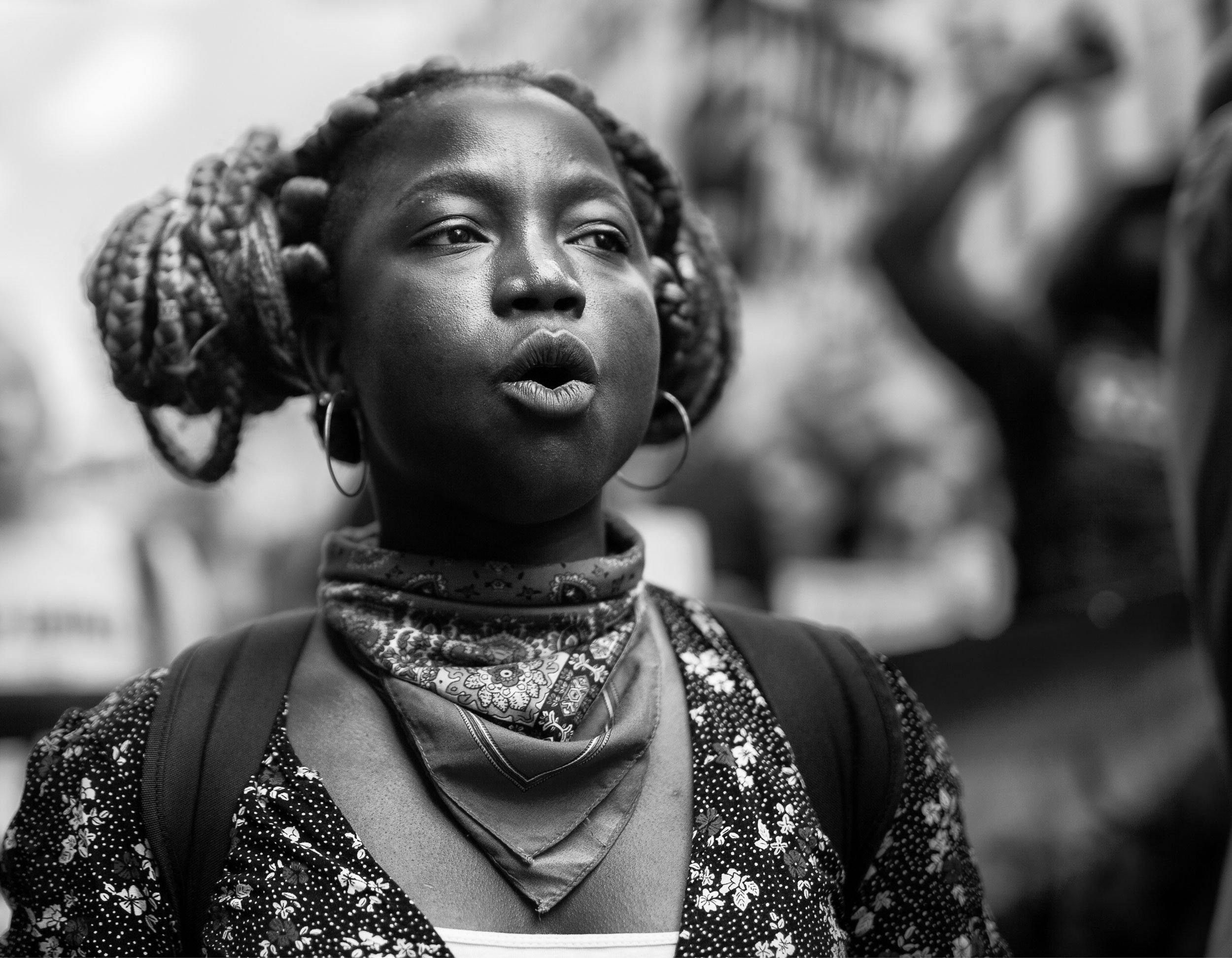 A photo of a woman with braided hair, wearing a patterned shirt, a bandana around her neck, and hoops in her ears. She has a backpack on and appears to be outdoors.