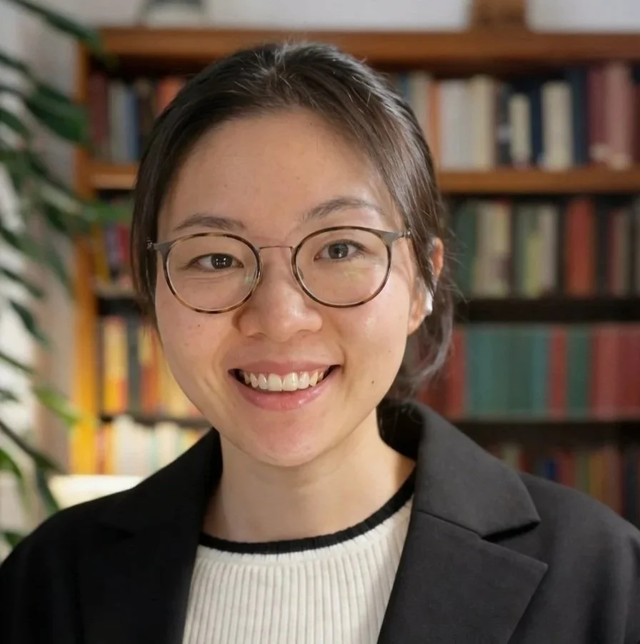A young woman with short brown hair, glasses, and a black blazer smiling in front of a bookshelf filled with books.