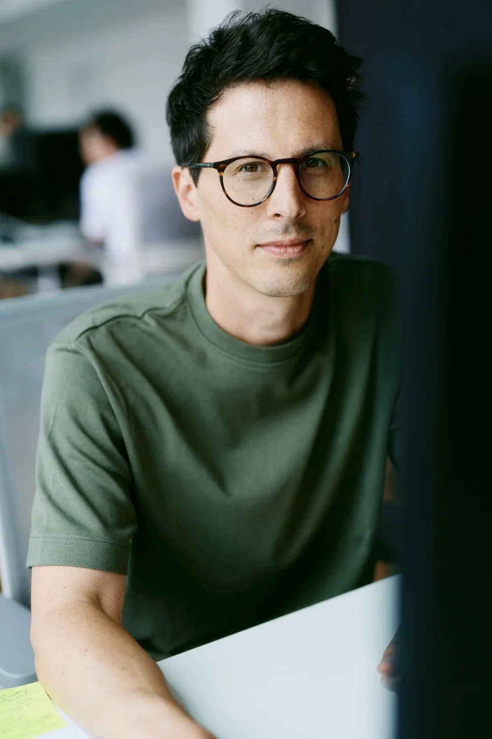 Ein junger Mann mit dunklem Haar, Brille und grüner T-Shirt sitzt an einem Schreibtisch in einem Büro, in die Kamera schauend.