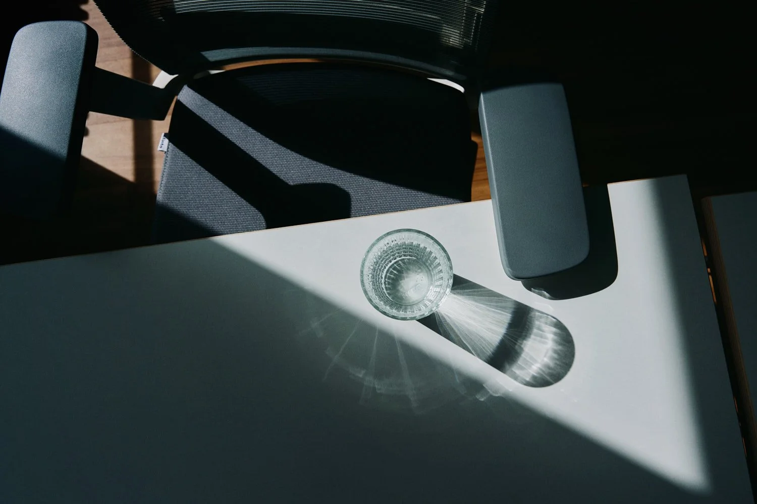 Ein Glas Wasser auf einem weißen Tisch, das Licht wirft einen Schatten und Lichtstrahlen auf den Tisch. Im Hintergrund ein Stuhl mit dunklem Sitz und Rückenlehne, auf dem Tisch steht nur das Glas Wasser.