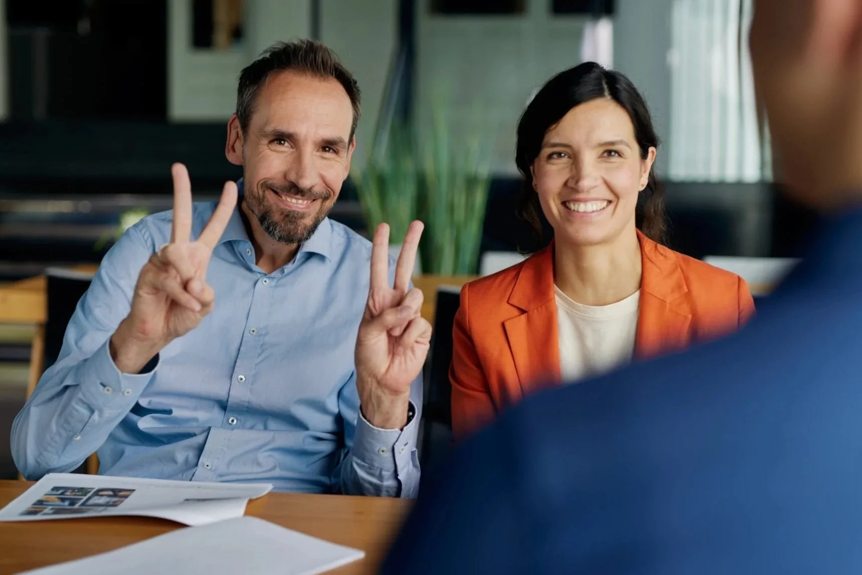 Zwei lächelnde Personen, ein Mann und eine Frau, zeigen peace-Zeichen in einem Büro. Der Mann trägt ein hellblaues Hemd, die Frau eine orange Jacke. Im Vordergrund ist eine Person im Gespräch, nur deren Schulter ist sichtbar.
