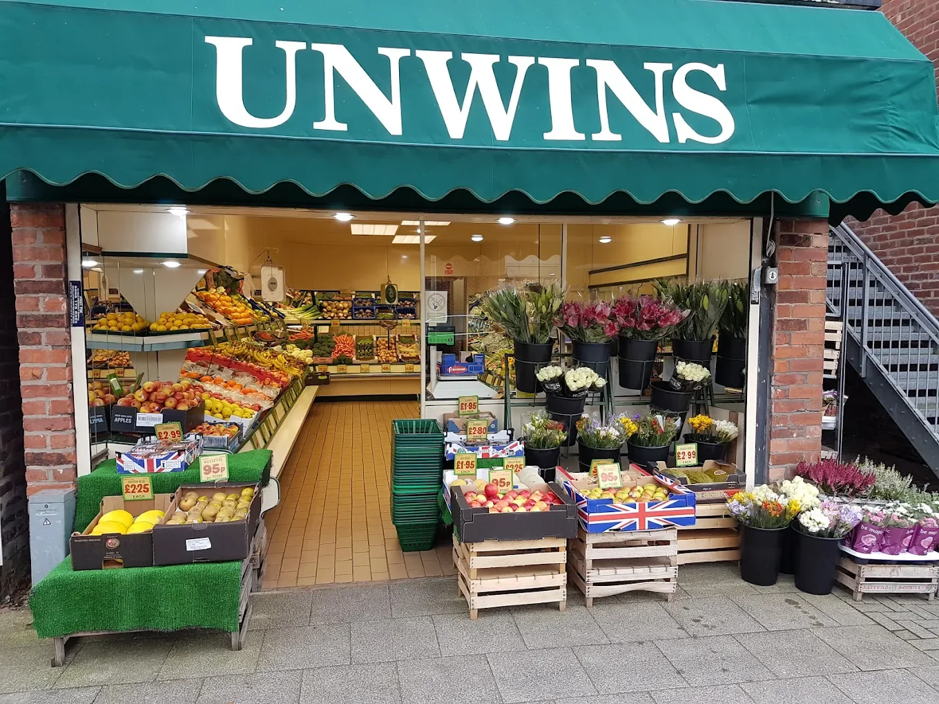 Front view of a small grocery store with a green awning labeled "UNWINS." The store displays various fruits like apples, bananas, and lemons, as well as flowers in black buckets and wooden crates outside.