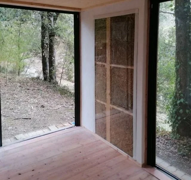 Interno di una stanza con pareti e pavimento in legno, con grandi finestre che mostrano un ambiente boschivo esterno con alberi e terreno naturale.