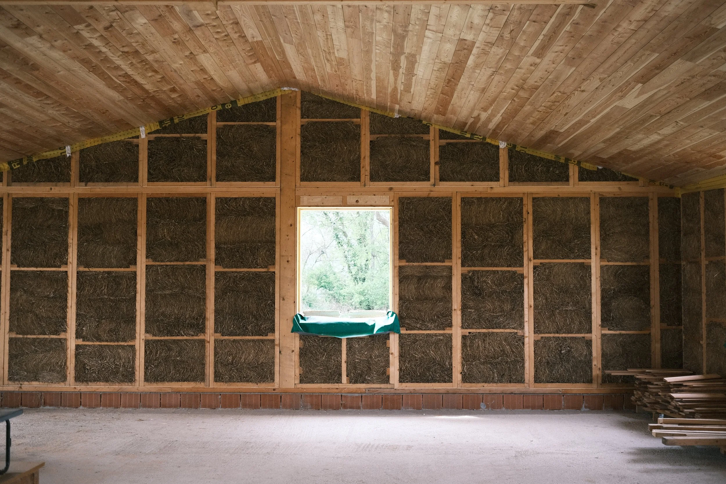 Interno di una stanza in costruzione con pareti di paglia e finestra, soffitto di pannelli di legno e pavimento di cemento.