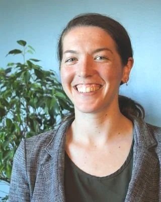 A smiling woman with dark hair tied back, wearing a gray blazer and a black shirt, standing next to a green plant against a blue wall.