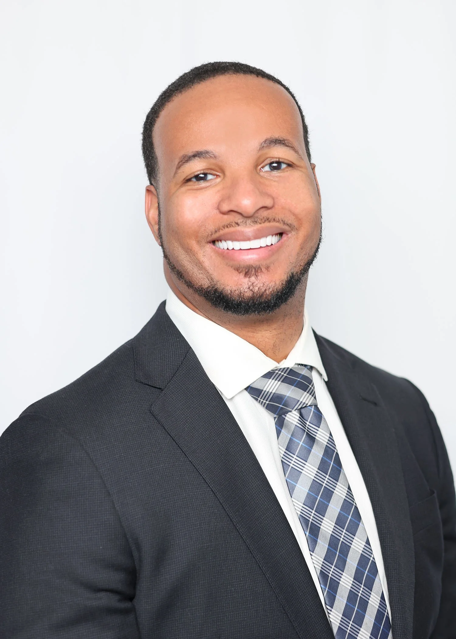 Professional headshot of a smiling man wearing a dark suit, white shirt, and plaid tie against a plain white background.