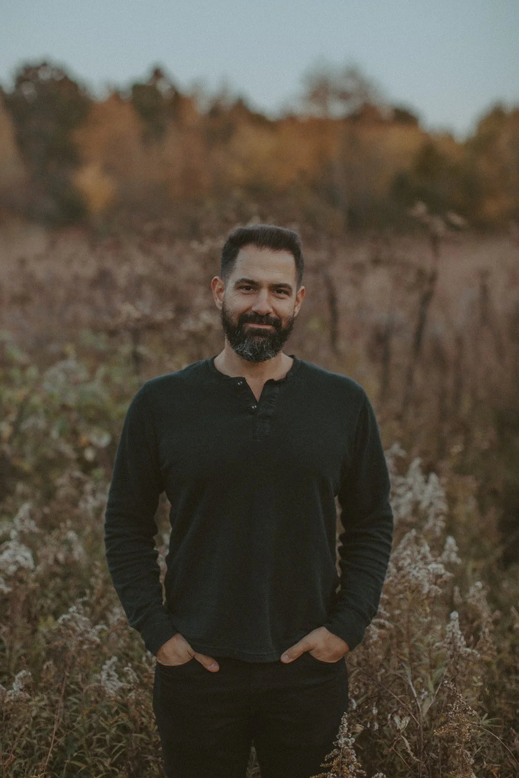 A bearded man in a black long-sleeve shirt standing outdoors with hand in pockets, a blurred hillside with trees in the background during a sunset.