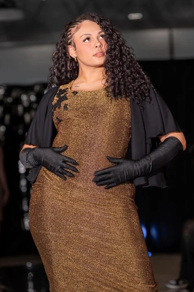 Woman with curly hair wearing a form-fitting gold dress, black gloves, and a black shawl, looking upward with her hands positioned on her waist.