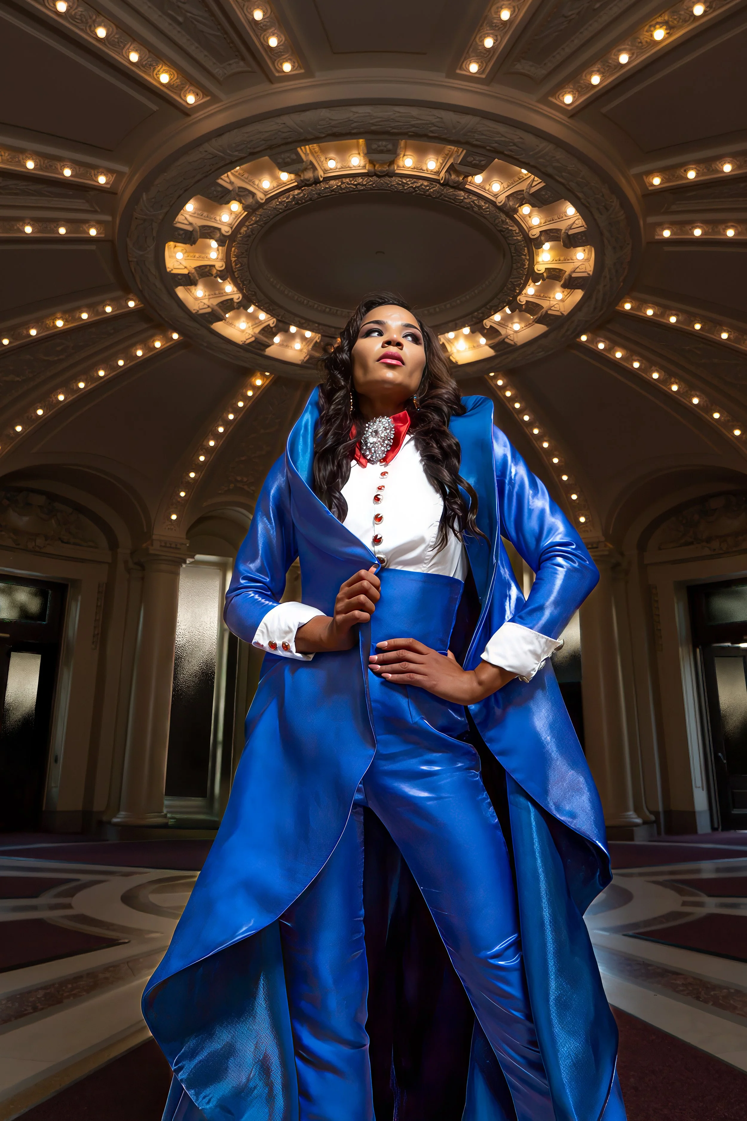 A woman in a shiny blue suit with white cuffs and red buttons poses confidently inside an ornate, well-lit interior with a large circular chandelier overhead.