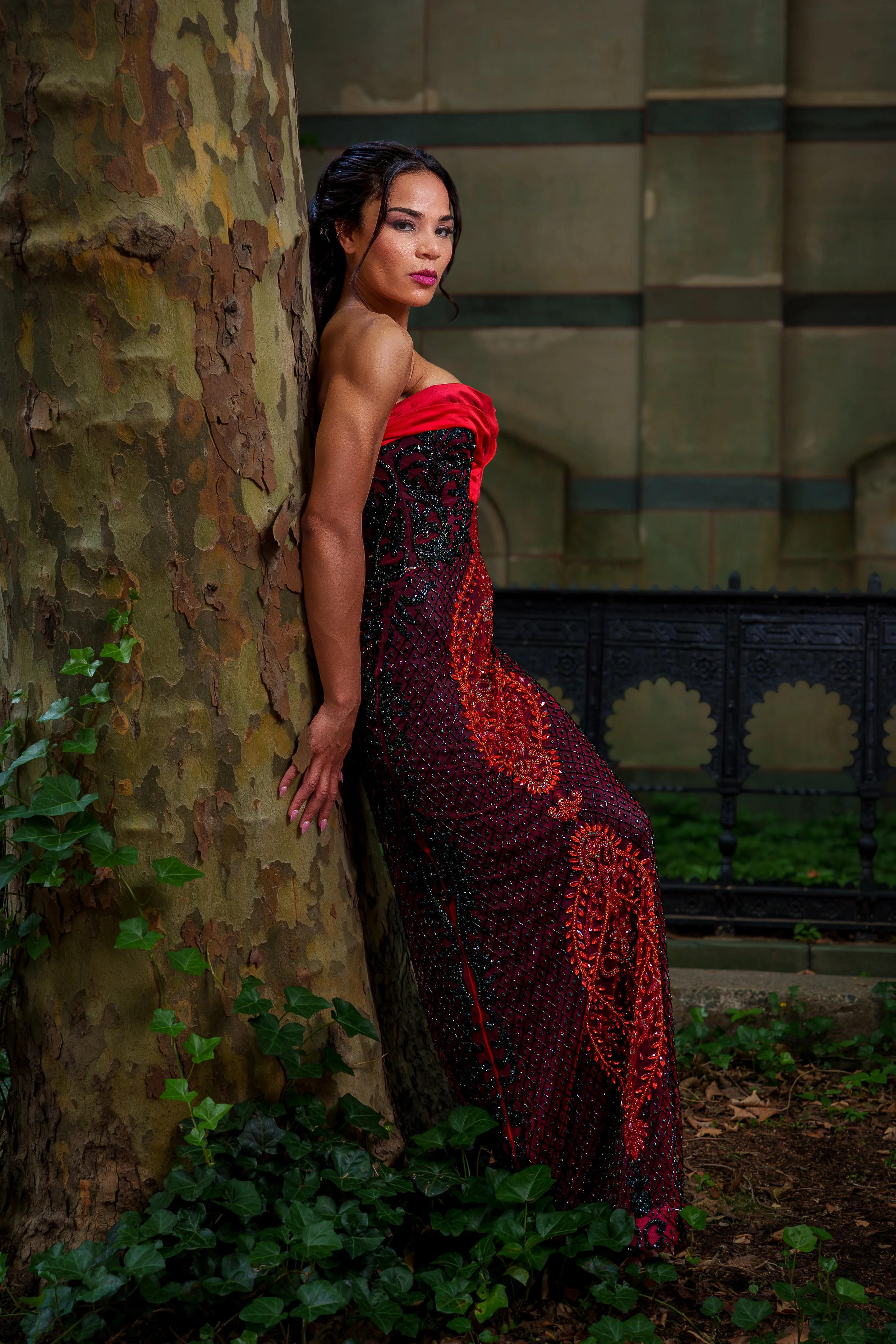 A woman in a red and black ornate dress standing behind a large tree with green ivy leaves on the ground, in an outdoor setting with a decorative black metal fence and a stone wall in the background.