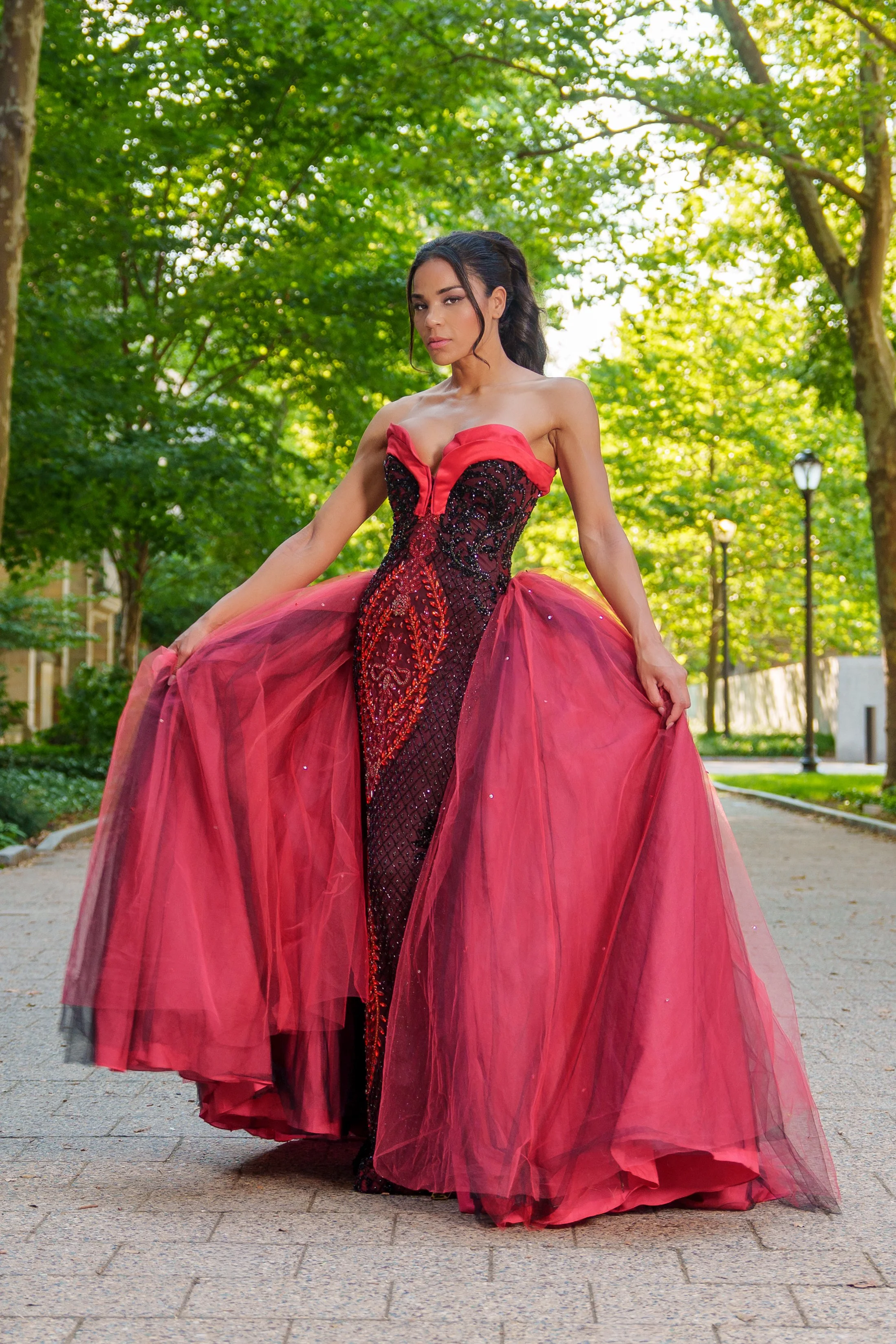 A woman in an elegant red and black strapless gown with a tulle skirt stands outdoors in a park with green trees and sunlight filtering through the leaves.
