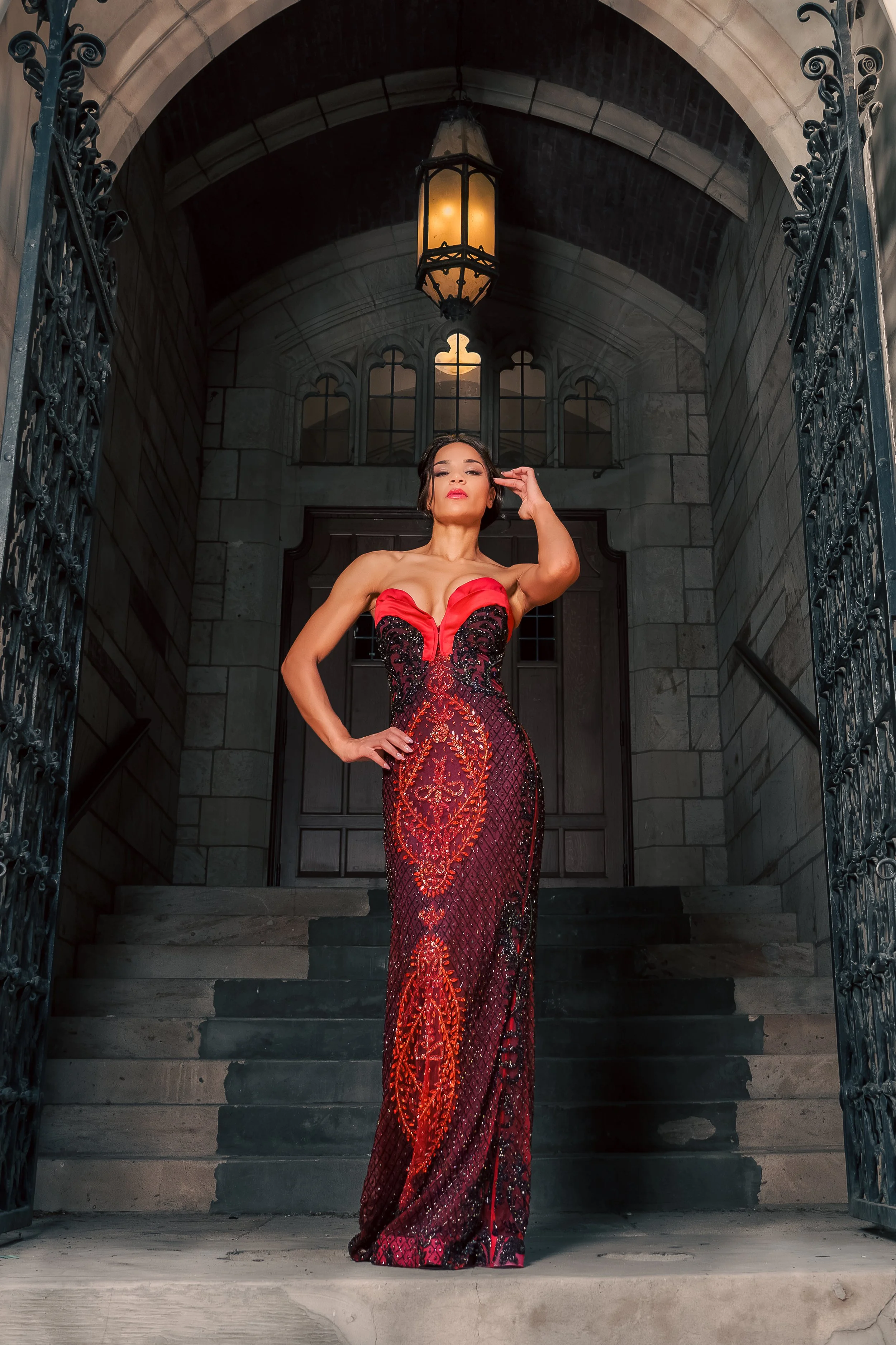 A woman in a red and black patterned evening gown with a deep neckline standing on stone steps beneath an arched stone doorway with wrought iron gates, a lantern hanging above.