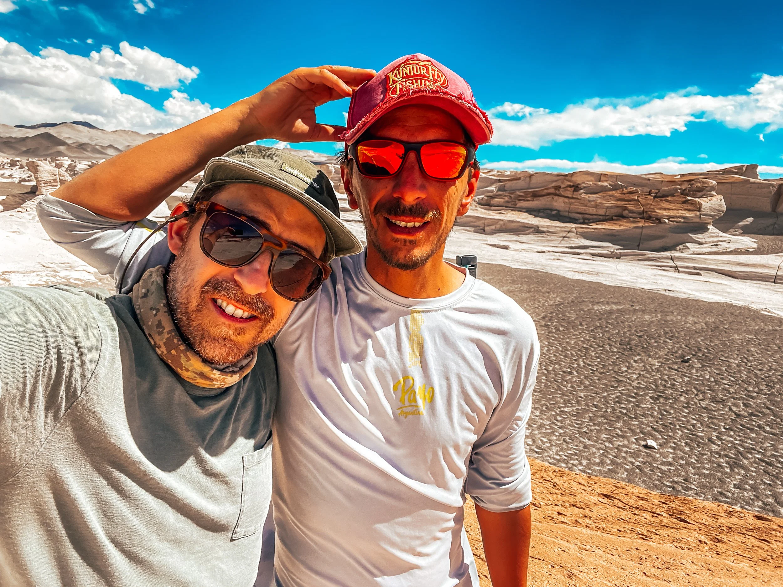 Two men take a selfie in a desert landscape with rock formations and mountains in the background, wearing hats and sunglasses, under a bright blue sky with scattered clouds.