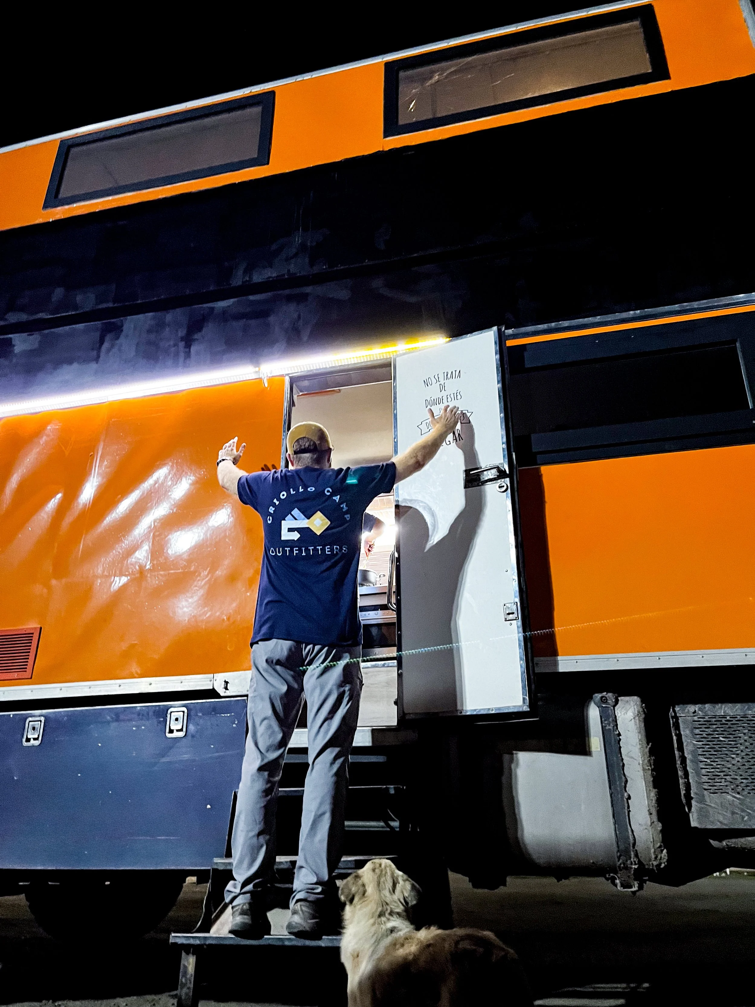 A man standing on a small staircase opening the door of a large orange bus at night, with a dog sitting on the ground in front of him.