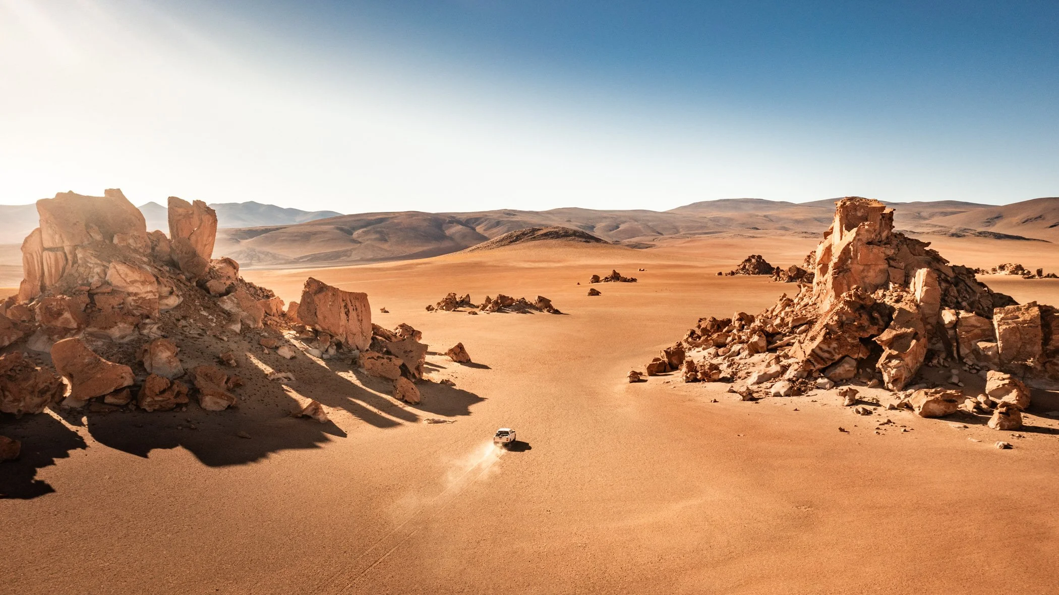A vast high-altitude desert landscape with large rock formations on either side, an off-road vehicle is driving across the sandy terrain, and distant mountains under a clear sky.