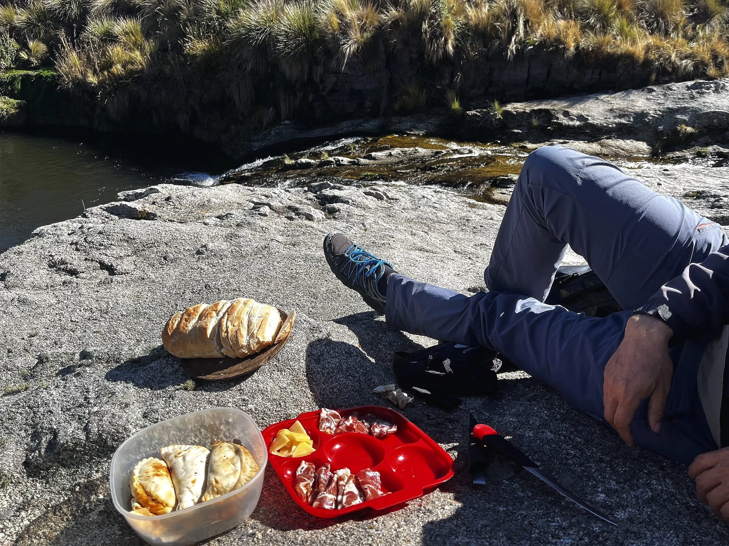 A person relaxing on a rocky riverbank with bread and snacks placed on rocks. The person is wearing gray outdoor clothing and blue hiking shoes, with a backdrop of water and grassy vegetation.