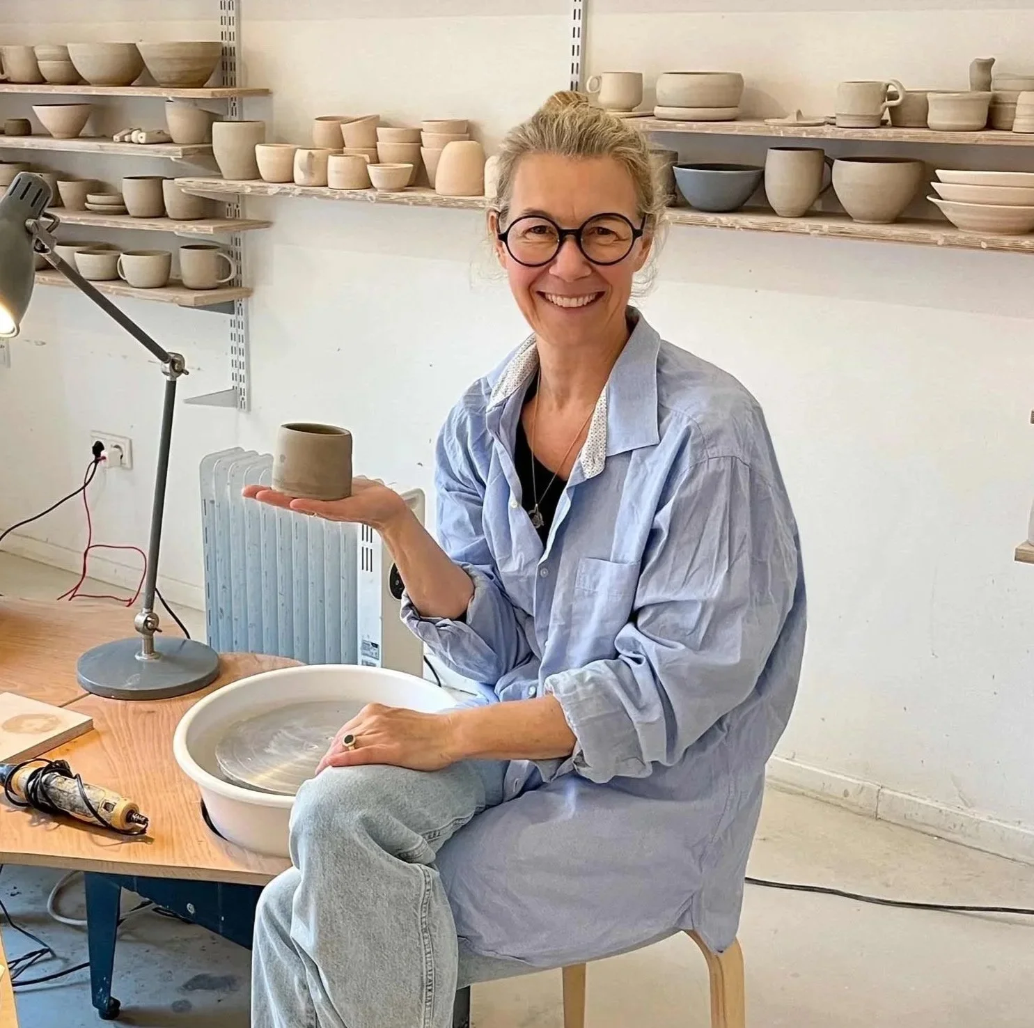 Eine Frau sitzt in einem Töpferstudio und zeigt stolz eine lederharte, selbstgetöpferte Tasse. Im Hintergrund sind Regale mit verschiedenen keramischen Objekten wie Tassen, Schalen, Becher zu sehen.